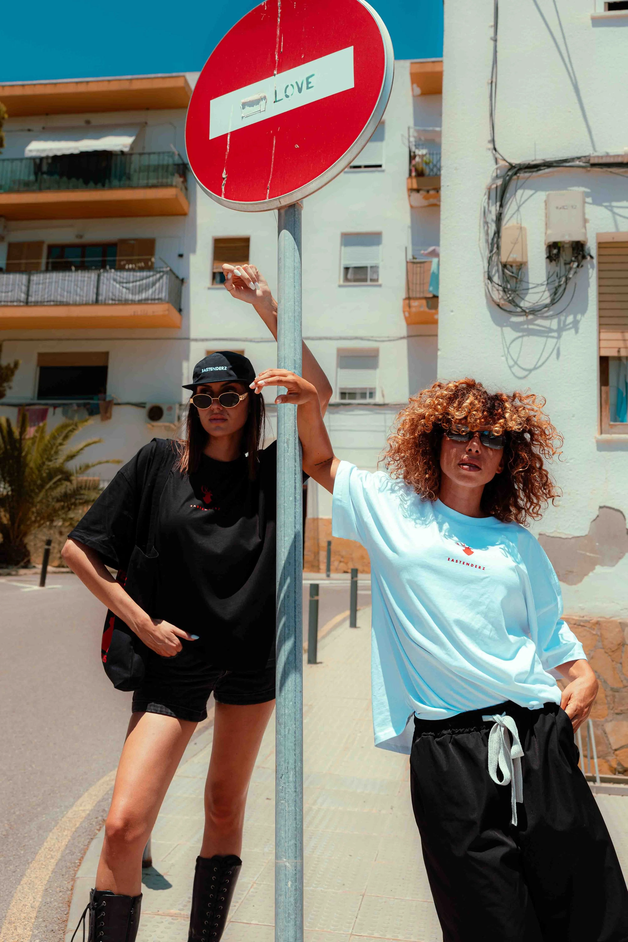 Two women standing near a street sign with the word "LOVE" on it. One woman has curly hair, sunglasses, and is wearing a white T-shirt and black pants. The other woman has long dark hair, sunglasses, a black cap, and a black T-shirt. They are outdoor