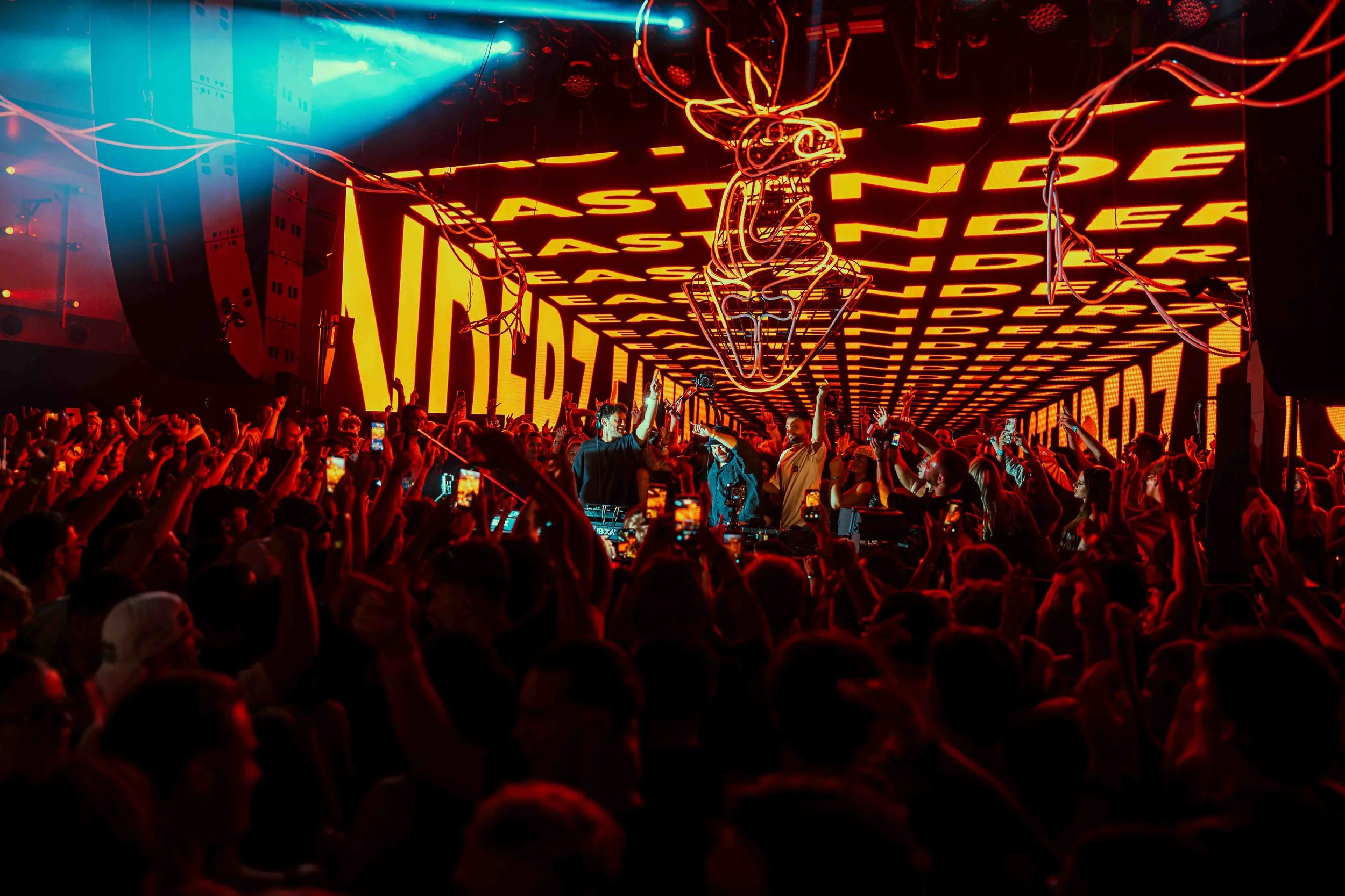A large crowd at a concert or nightclub with bright red and yellow neon lights and digital screens displaying the words 'EASE' repeatedly. A DJ or performer is visible in the center with equipment, and people are raising their hands and taking photos