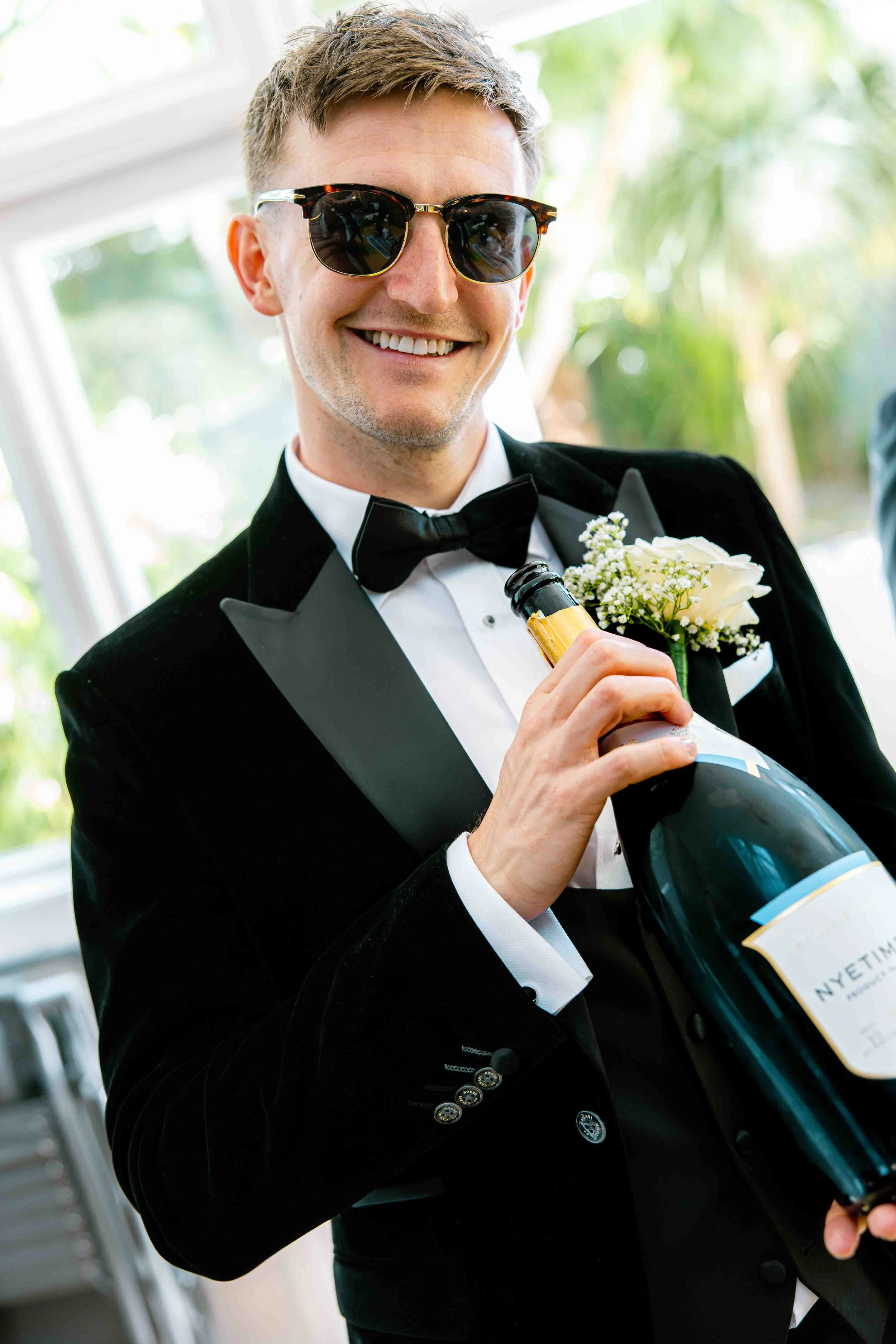 A man in a tuxedo and sunglasses holding a bottle of champagne, smiling.