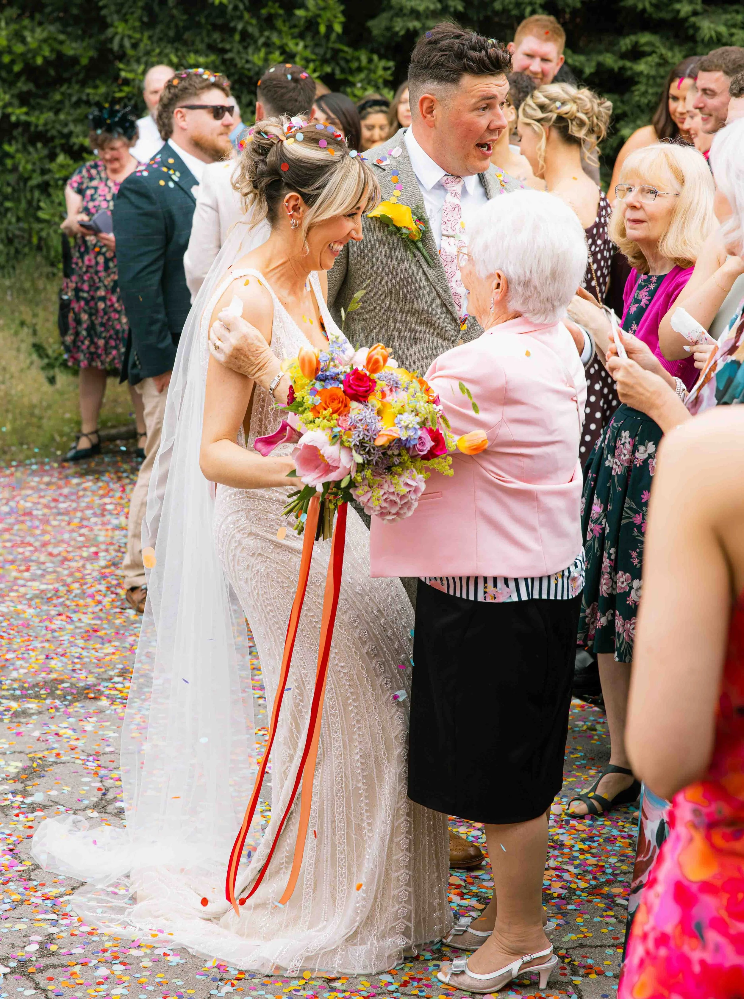 A wedding celebration outdoors with confetti on the ground. The bride, in a white lace wedding dress, is holding a bouquet of colorful flowers and is smiling while talking to an older woman wearing glasses and a pink jacket. The groom, in a gray suit