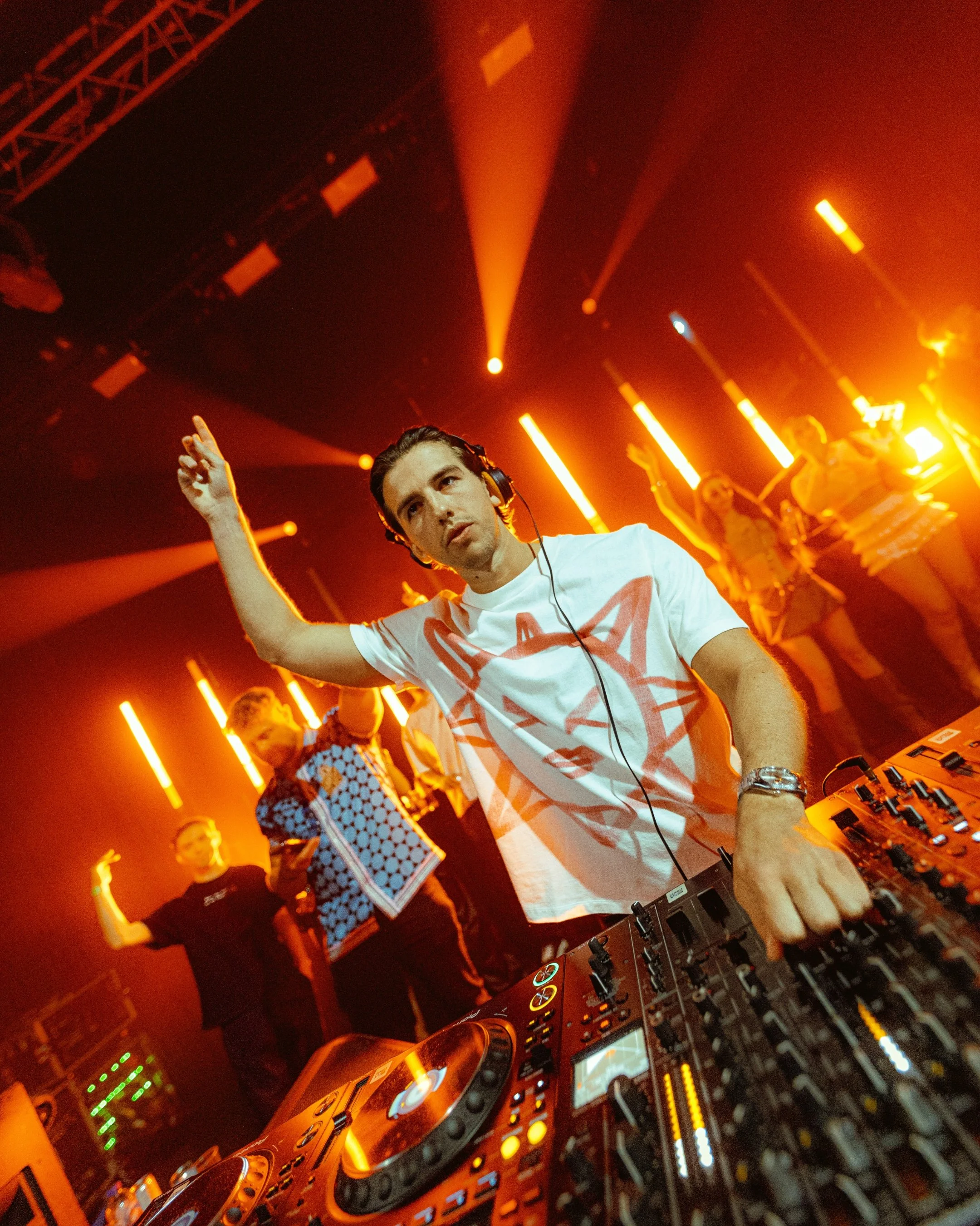 Josh baker with headphones standing behind a DJ mixer at a nightclub with orange lighting and people dancing in the background.