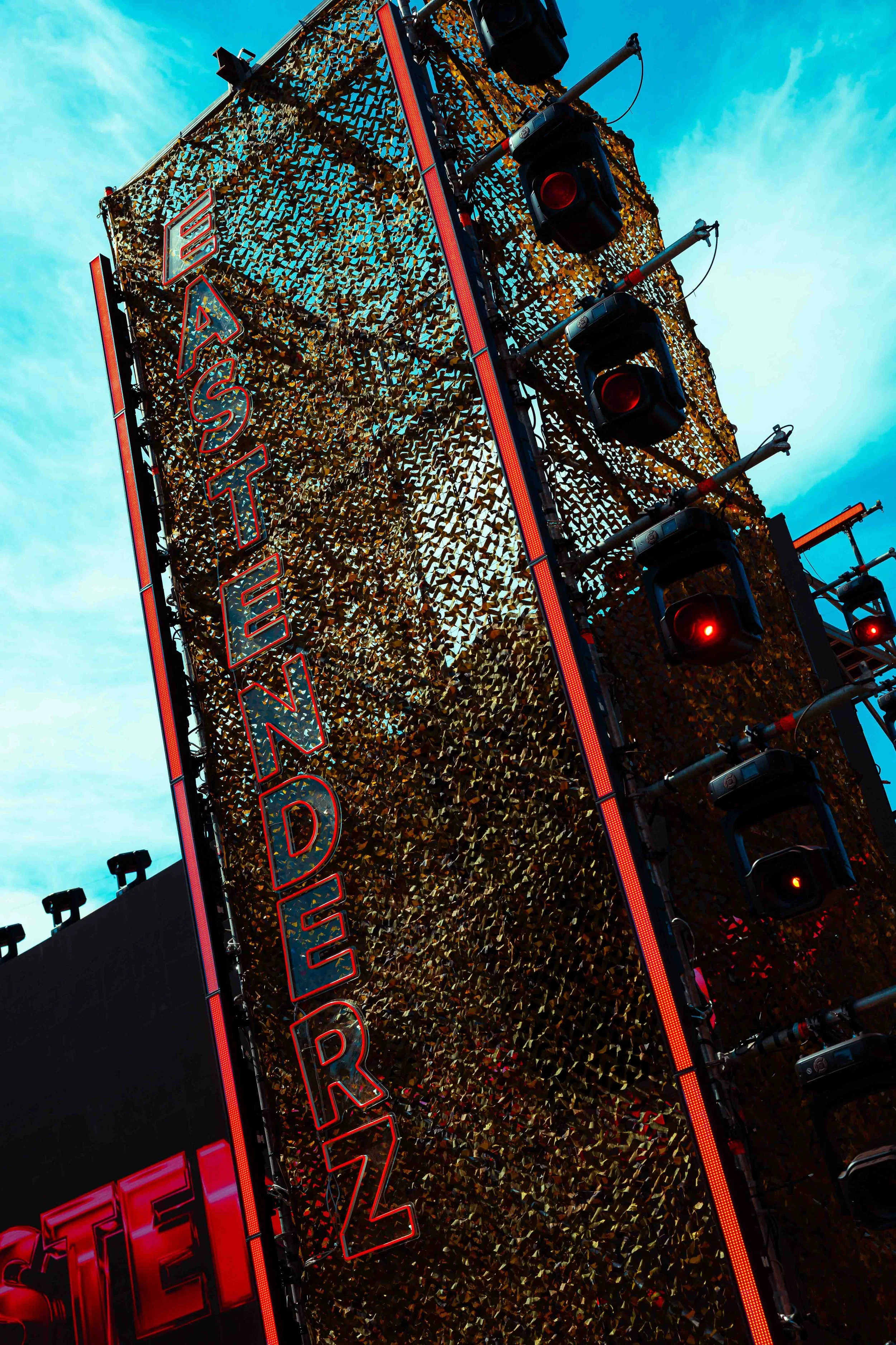 Brightly lit vertical sign with red neon outline reading 'EAT', against a background of a patterned gold surface, with traffic lights in front showing red lights, and blue sky with clouds above.