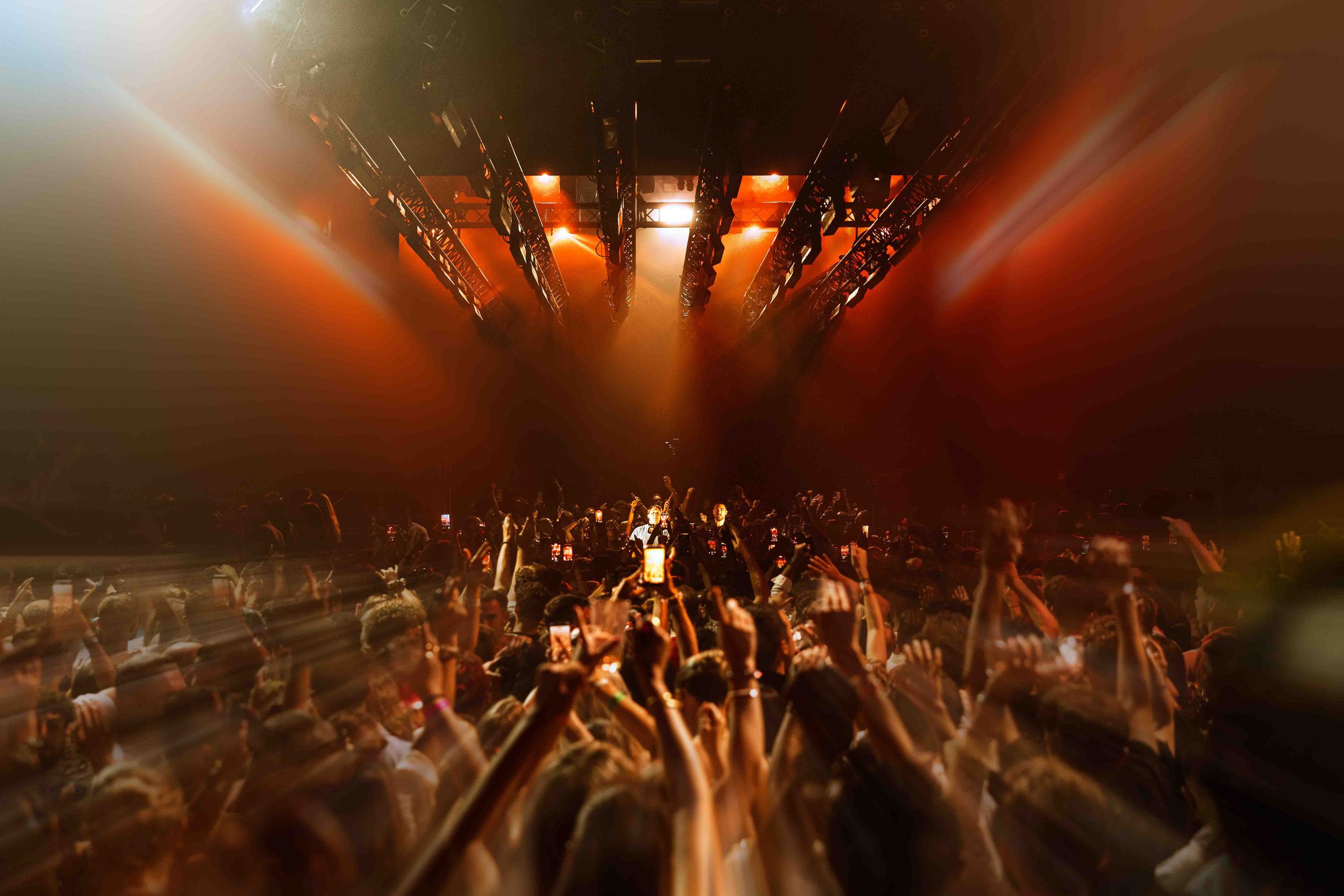 A concert at Hi ibiza with Camelphat, a large crowd of people taking photos and videos with their phones, illuminated by stage lights and beams of light shooting upwards towards a stage with bright, fiery lighting.