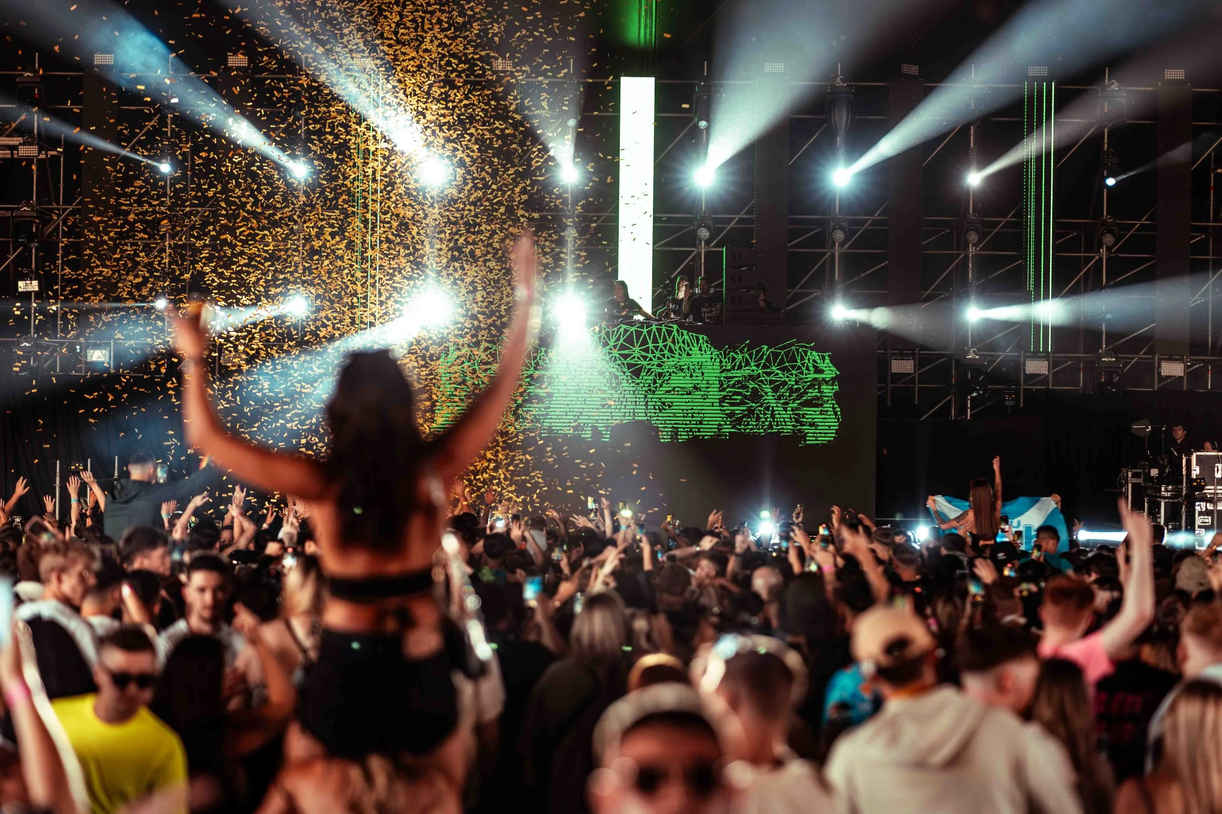 Crowd of people at a concert with hands raised, bright stage lights, confetti, and a DJ or performer on stage.