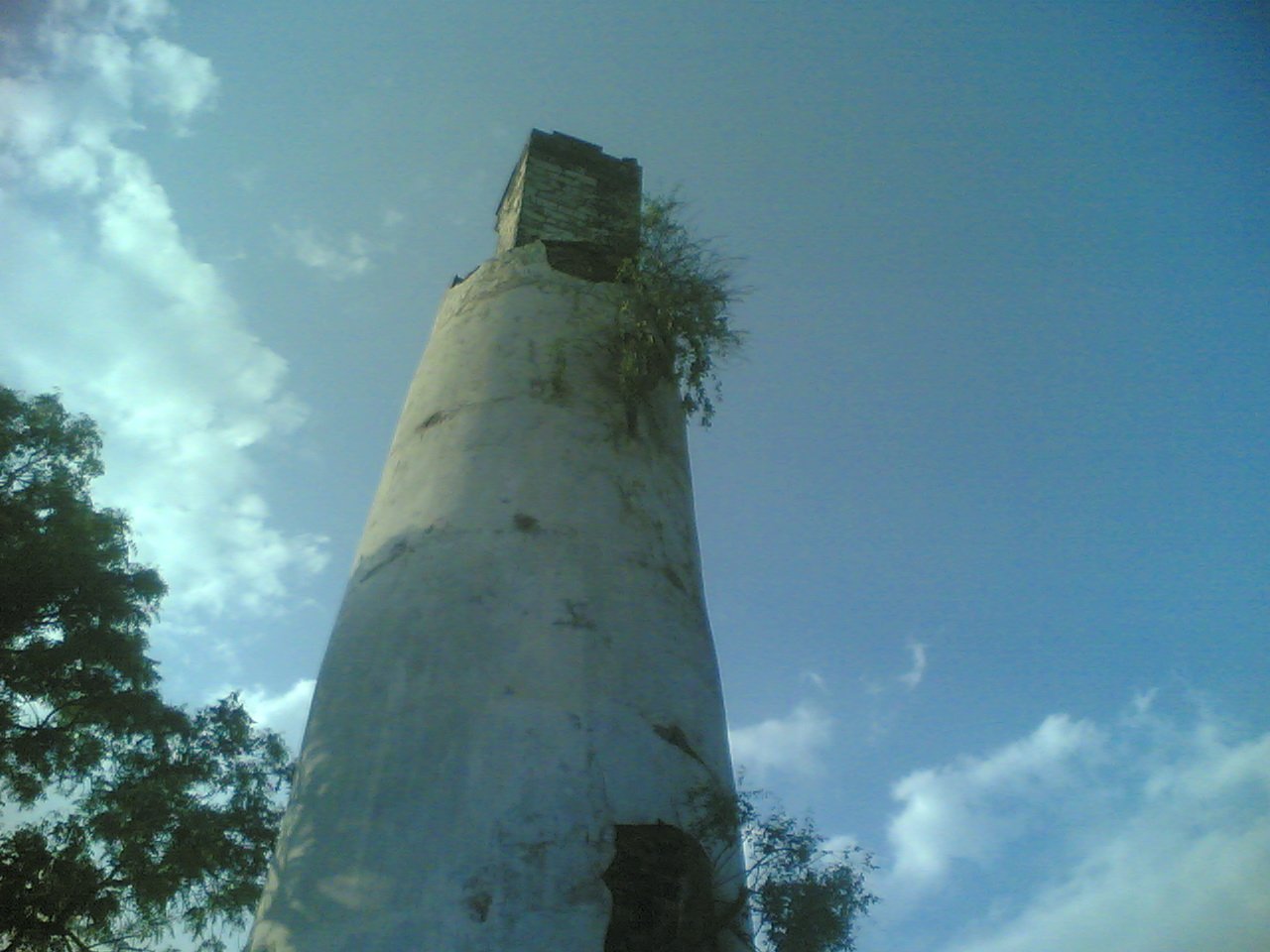 A tall, weathered white chimney or tower with a small wooden structure at the top, surrounded by trees and a blue sky with clouds.