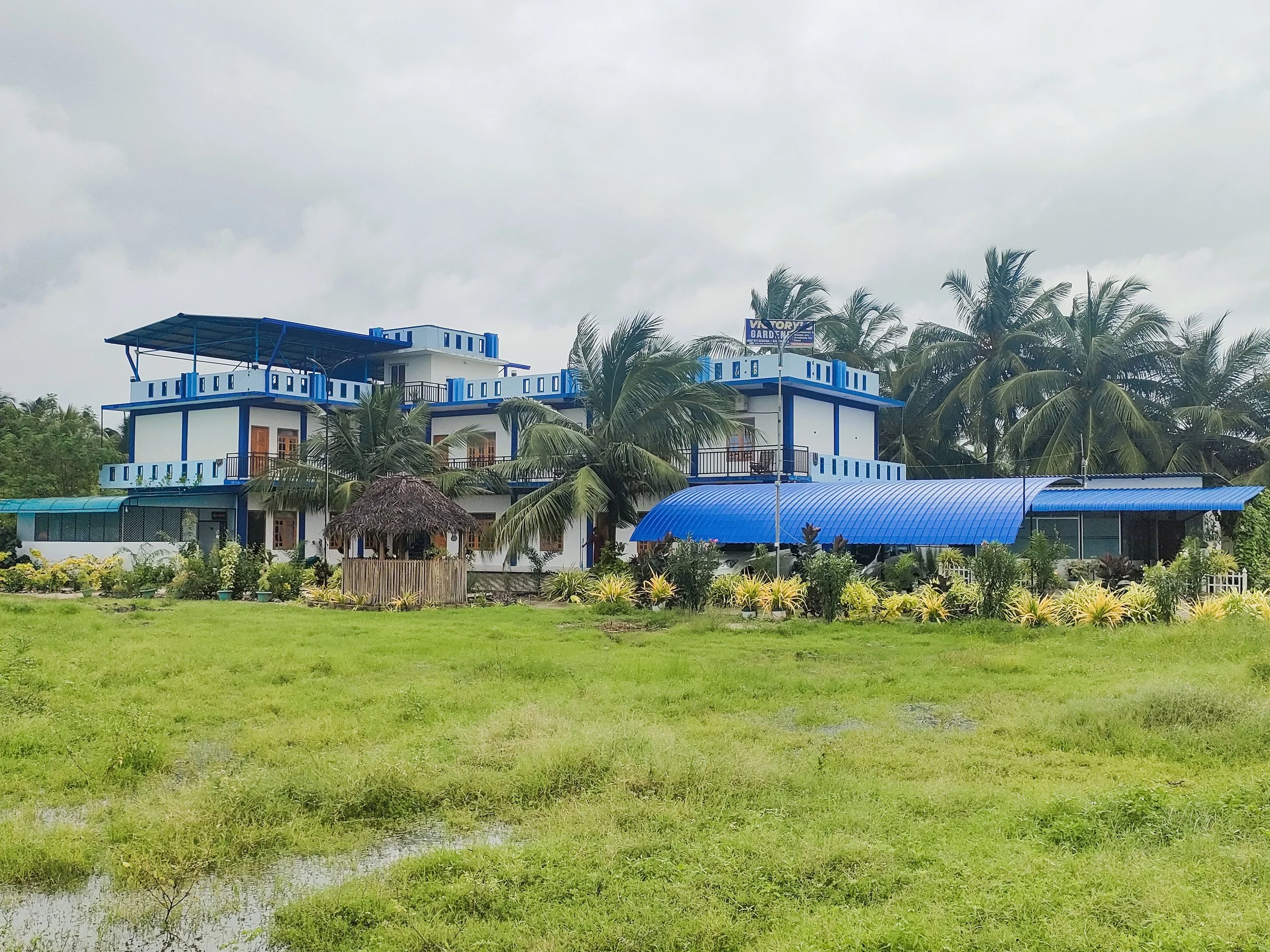 A multi-story building painted in white and blue, with balconies, surrounded by palm trees and lush green vegetation under an overcast sky.