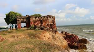 Ruins of an old brick building on a grassy cliff overlooking the ocean, with trees and clouds in the background.