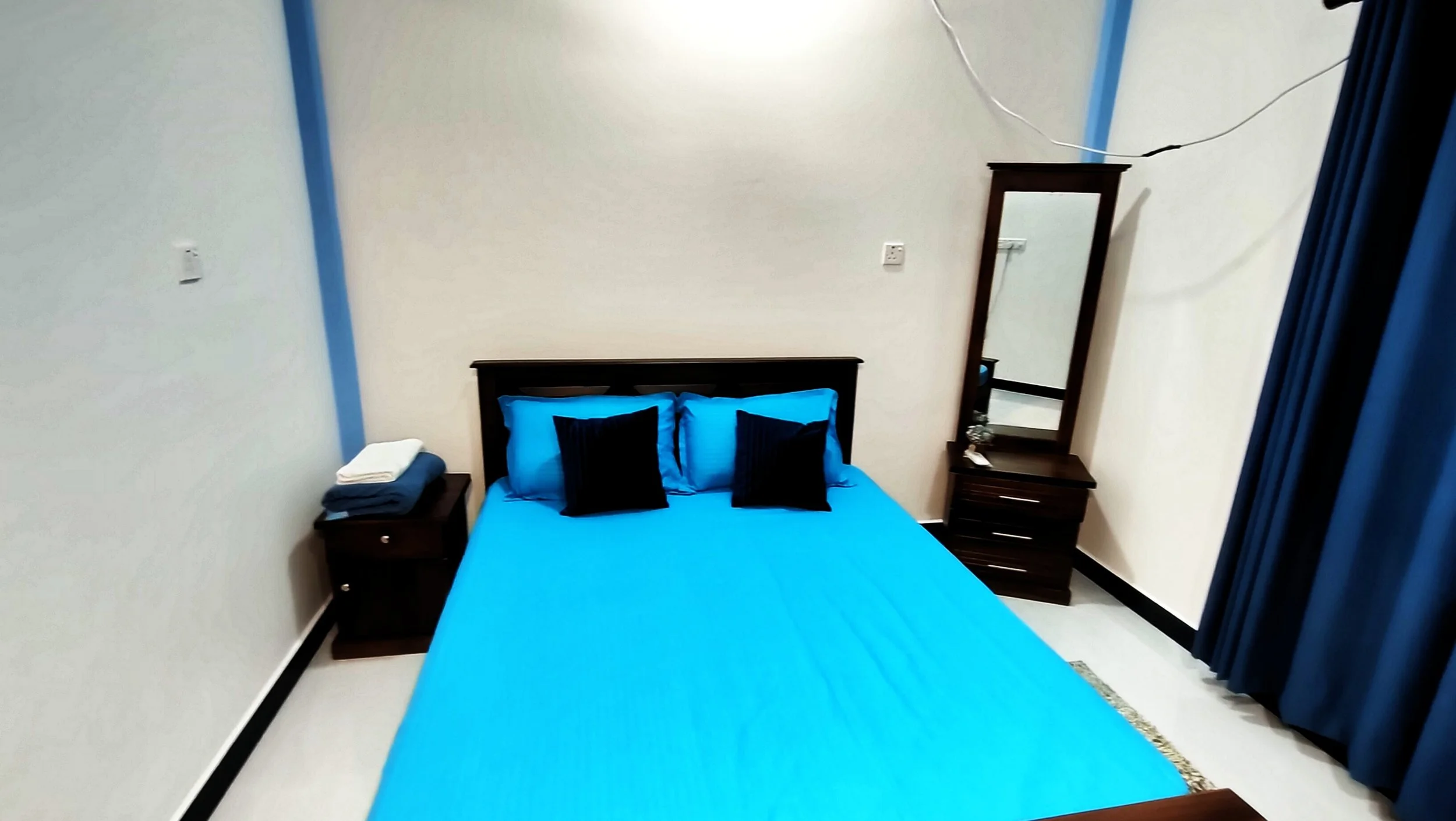 A bedroom with a bed dressed in blue sheets and black pillows, a wooden nightstand with folded towels, a tall mirror, and blue curtains.