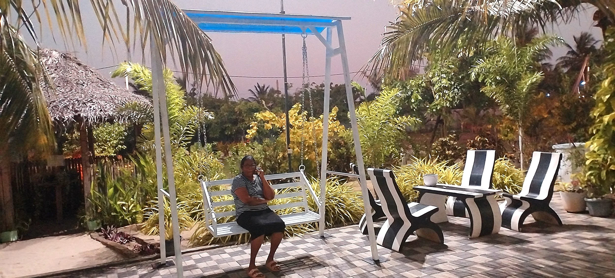 A woman sitting on a white garden swing in a backyard with lush green foliage, a thatched roof structure on the left, and black and white striped furniture, all under a partly cloudy sky