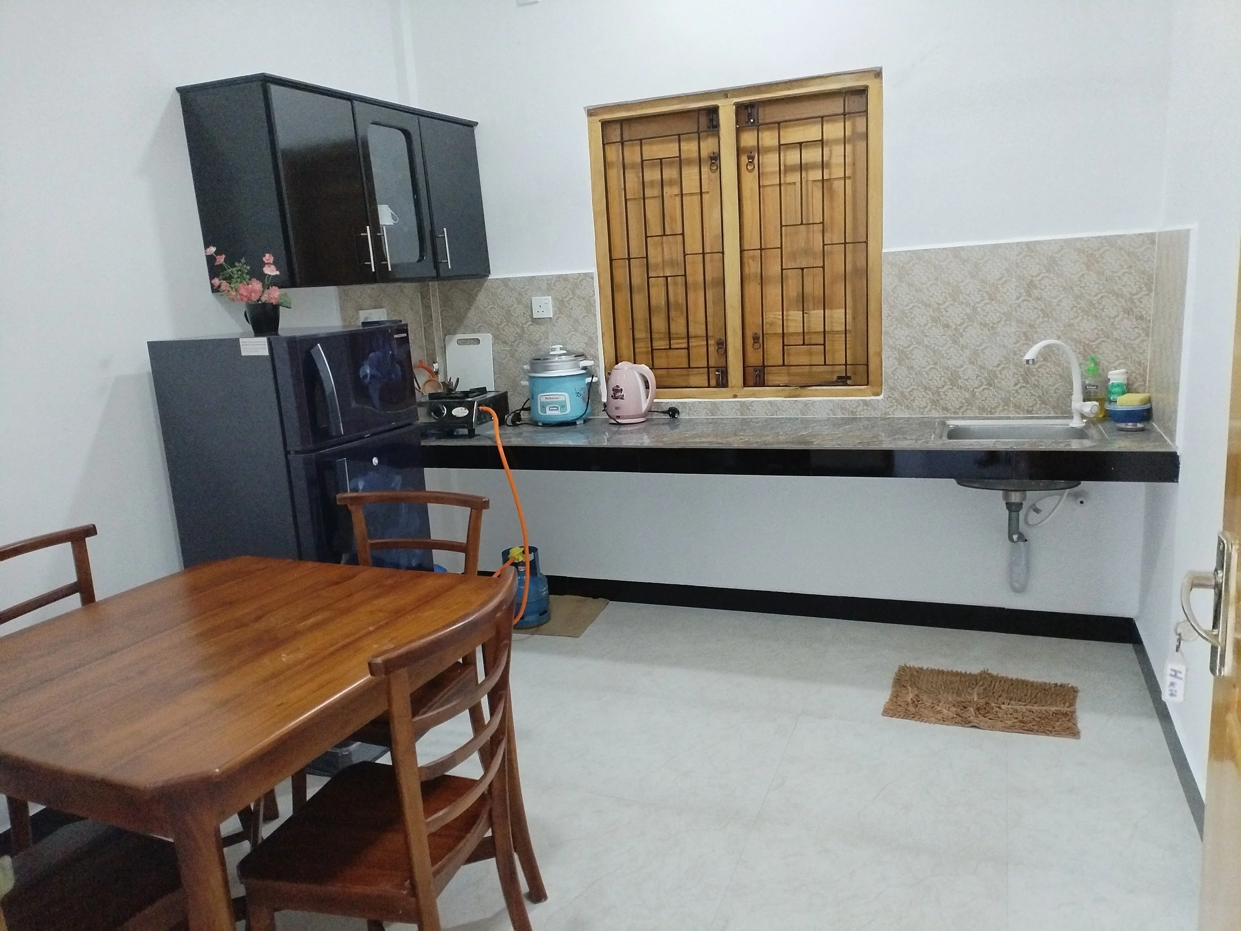 A kitchen with black cabinets, a gray countertop, a small sink, and a window with wooden shutters. There is a pink kettle, rice cooker, and some kitchen supplies on the countertop. A wooden dining table with chairs is in the foreground.