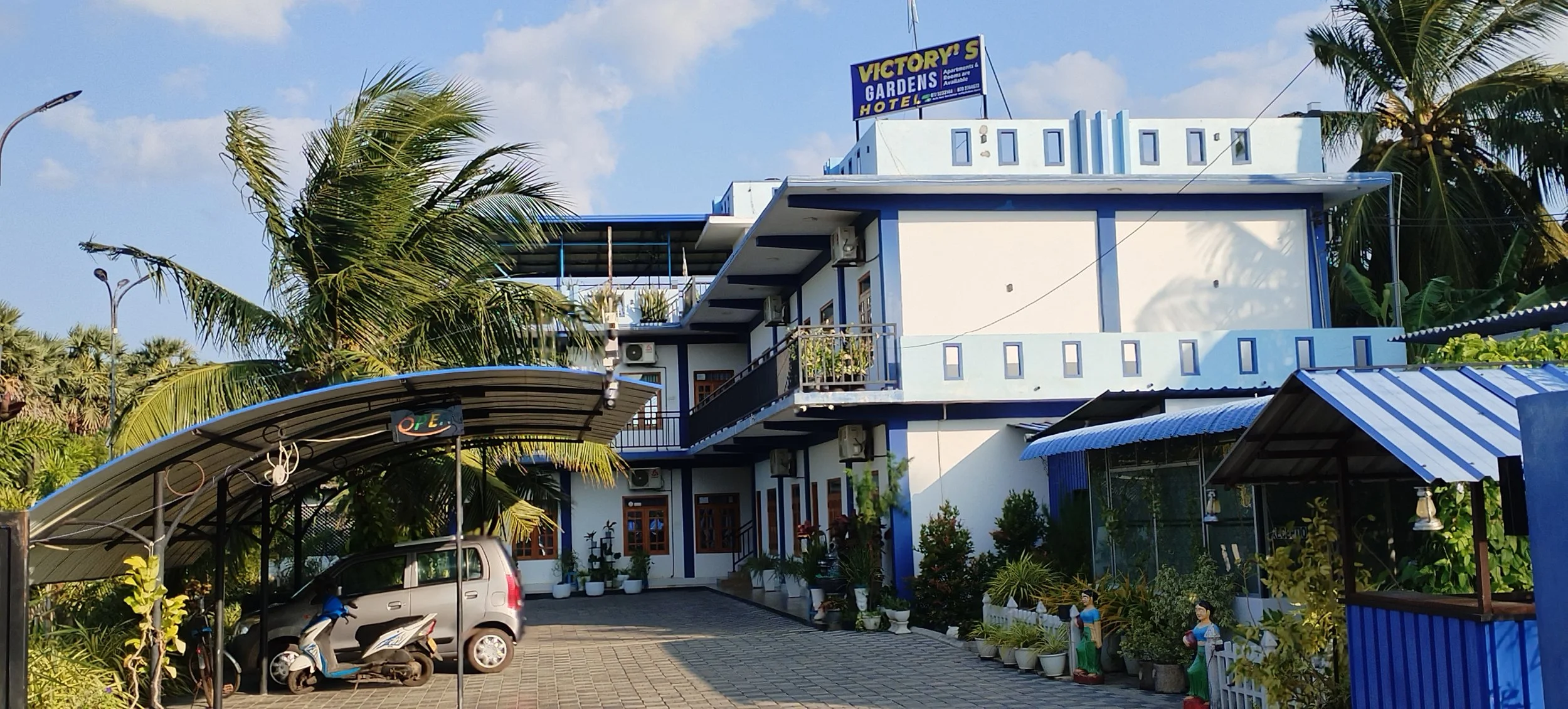 A white building with blue accents, featuring a balcony, multiple air conditioning units, and potted plants outside. There is a parking area with a car and two scooters and a covered bike parking area. Tall palm trees are surrounding the building, and a sign on the roof reads 'Victory's Gardens Hotel.'