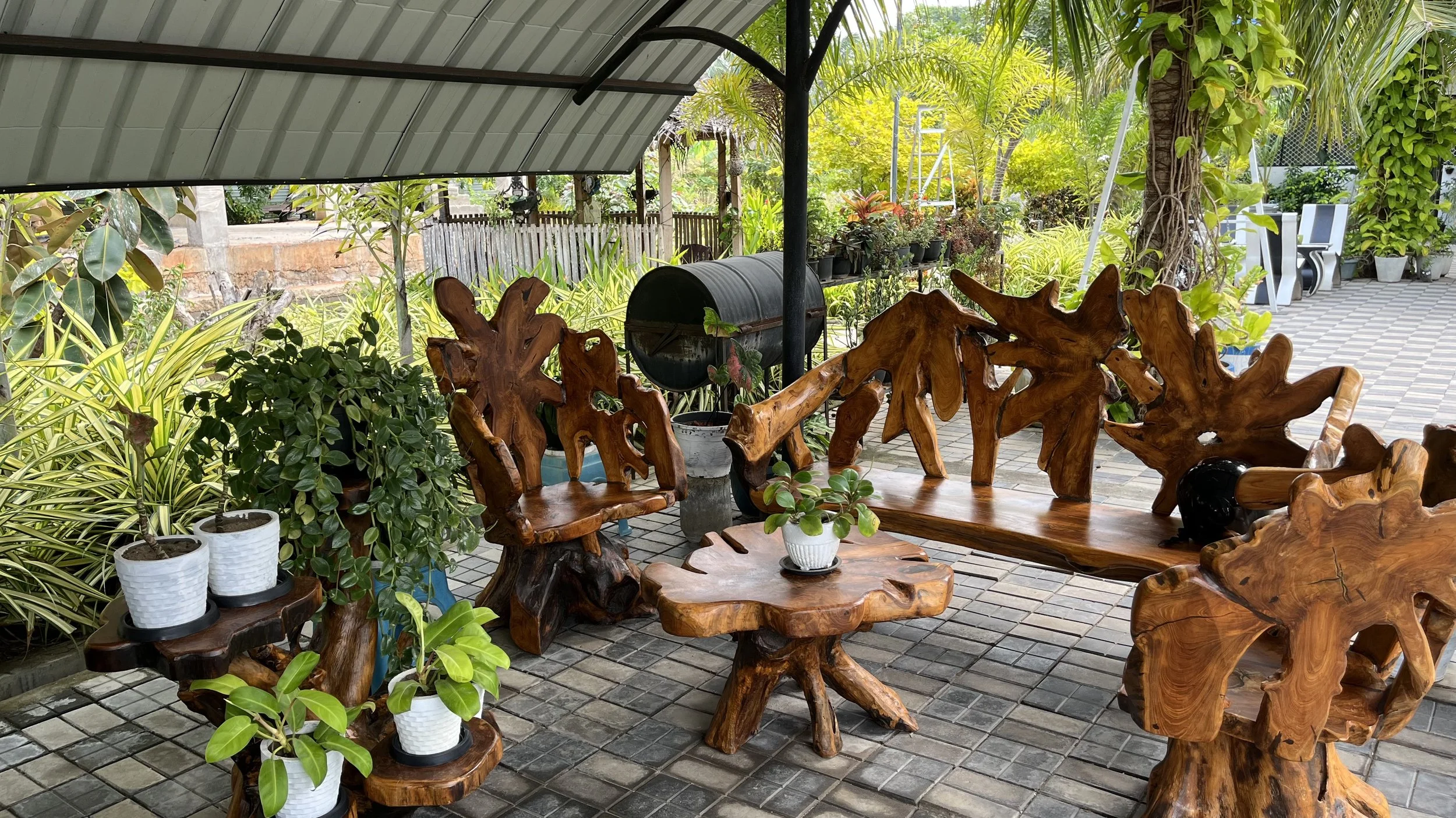 Outdoor patio with wooden furniture, potted plants, and greenery including trees and shrubs under a metal roof.