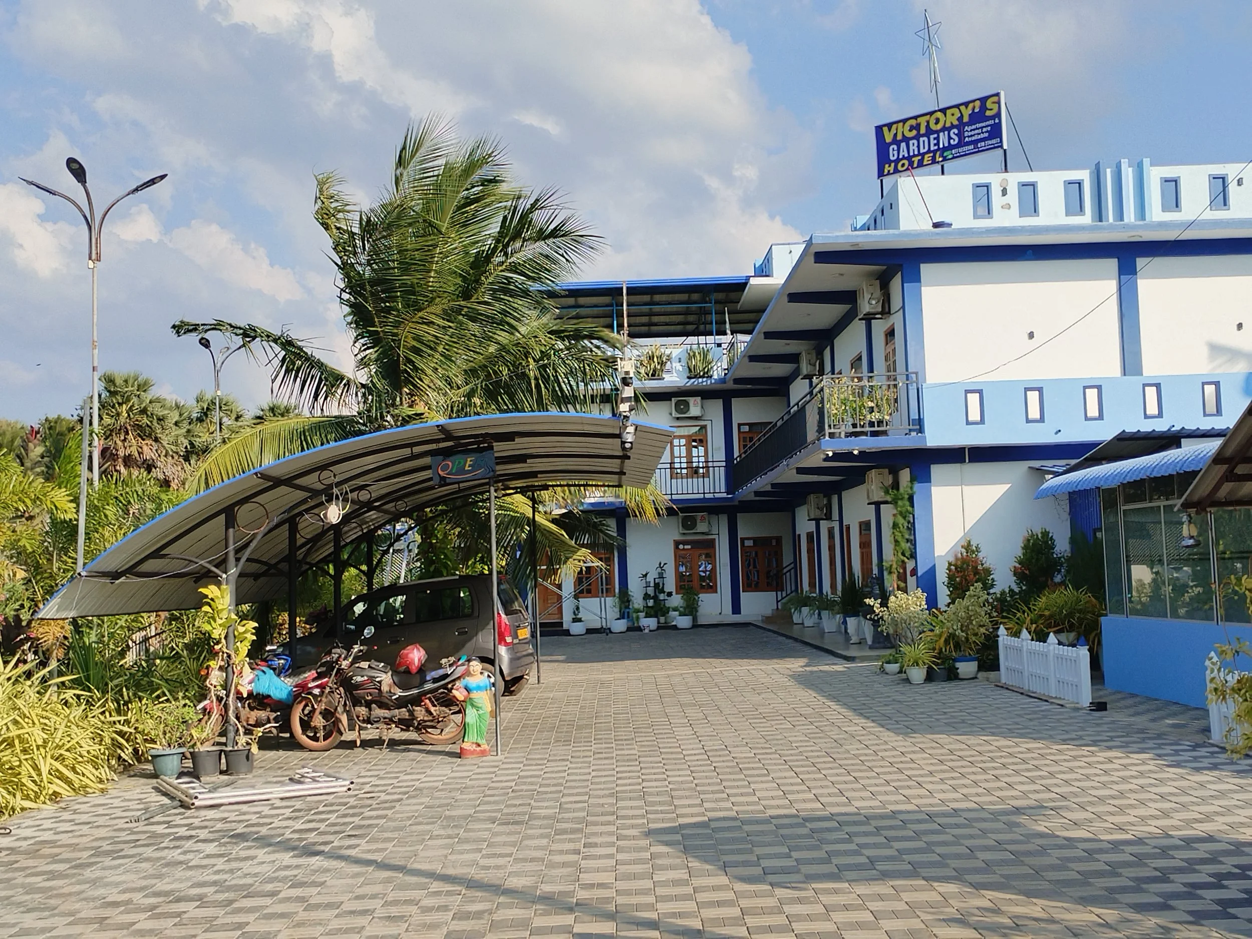 A blue and white building with a sign that says 'Victory's Gardens Hotel'. The parking area in front has motorbikes, a car, and a decorative garden statue of a woman. There are potted plants and a roofed structure with a sign that reads 'Open'. Tall 