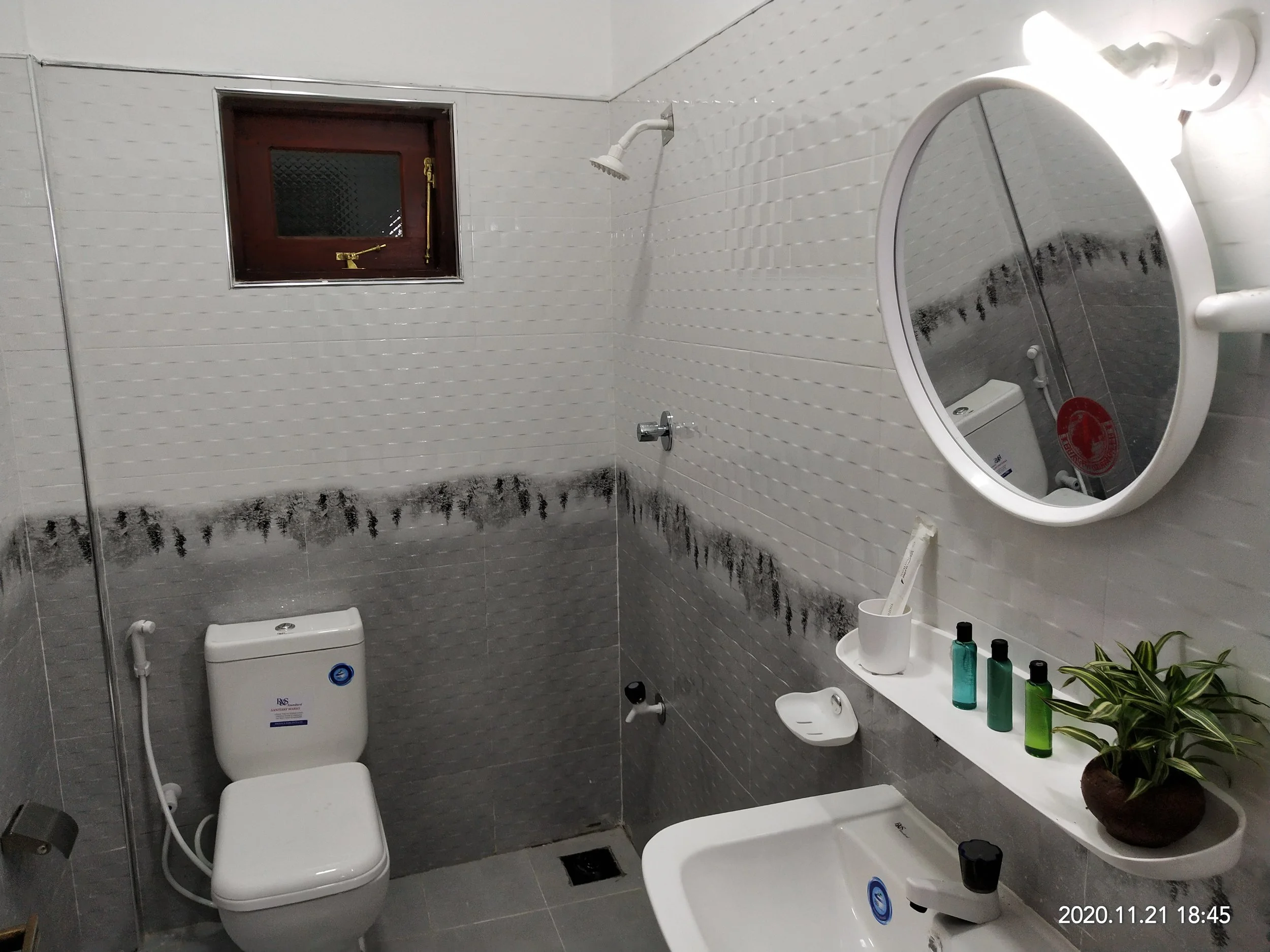 A bathroom with a wall-mounted showerhead, a small window with a dark brown frame, a white toilet, and a sink with a large mirror. There is a counter with several bottles of soap or shampoo and a potted plant. The walls are tiled with white tiles, an