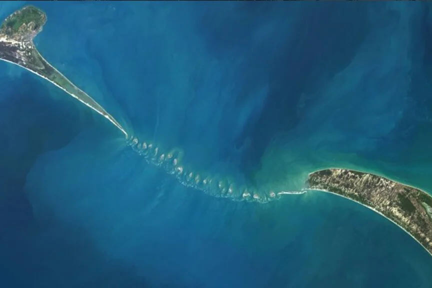 Aerial view of a barrier island and a causeway connecting it to the mainland, across a body of water with a series of small, rocky islands or sandbars in the water.