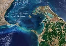 Aerial view of a coastal area with a narrow water channel between two landmasses of India and Sri Lanka, featuring lush greenery on the land and deep blue ocean water.