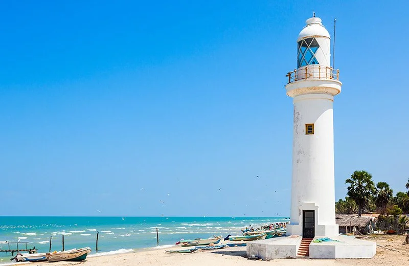 White lighthouse on a sandy beach with boats and palm trees, blue ocean and sky in the background.
