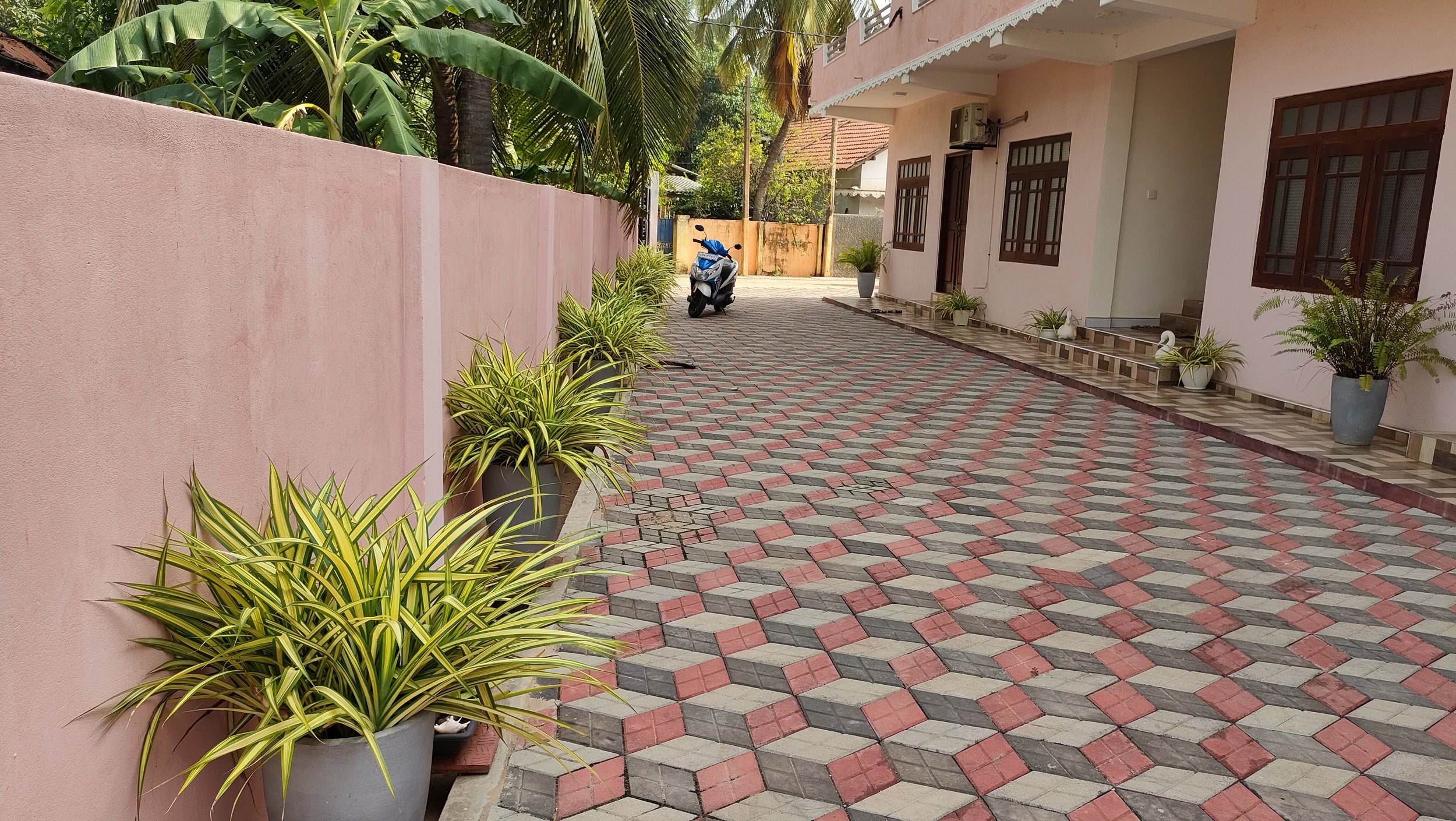 A paved driveway with red, gray, and beige interlocking tiles, lined with potted plants and a pink house wall on the left, a motorcycle parked near the house, and tropical trees in the background.