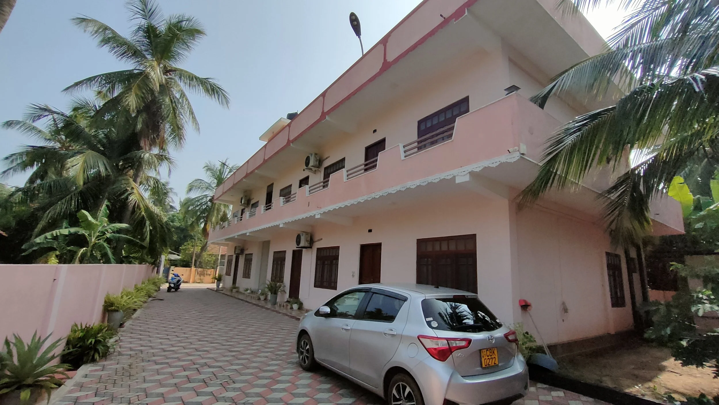 A three-story pink residential building with balconies, surrounded by palm trees. There are air conditioning units on the second and third floors. A grey car is parked in front, along with a scooter nearby. The sky is clear.