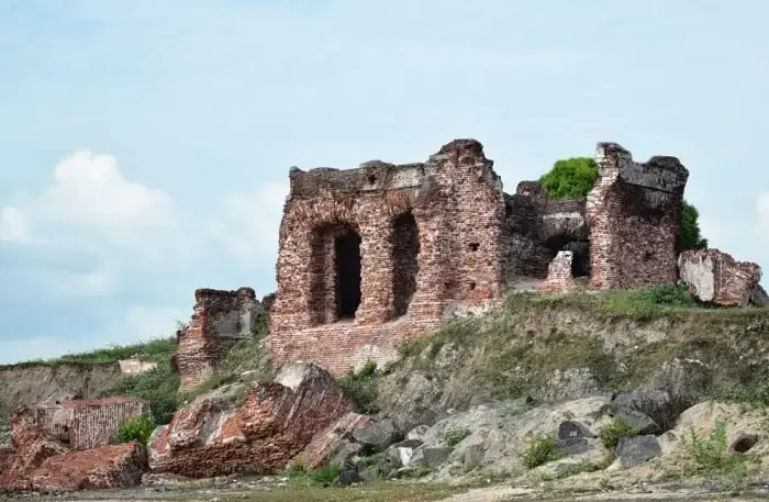 Ruins of an old brick building on a grassy hill with rock debris in the foreground, under a partly cloudy sky.
