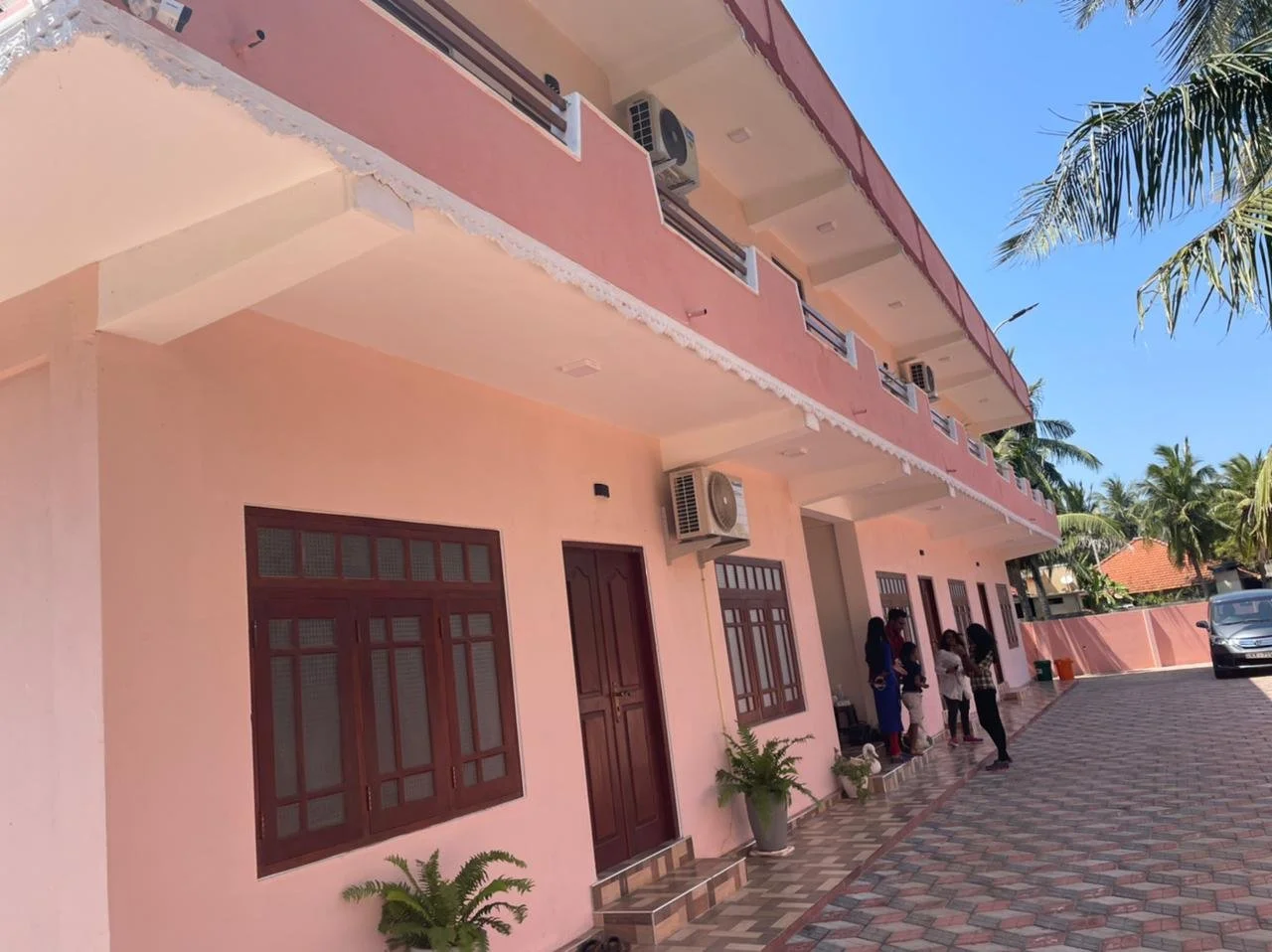 Pink two-story building with multiple windows and doors, surrounded by palm trees. A group of people is standing outside, and there is a white car parked nearby.