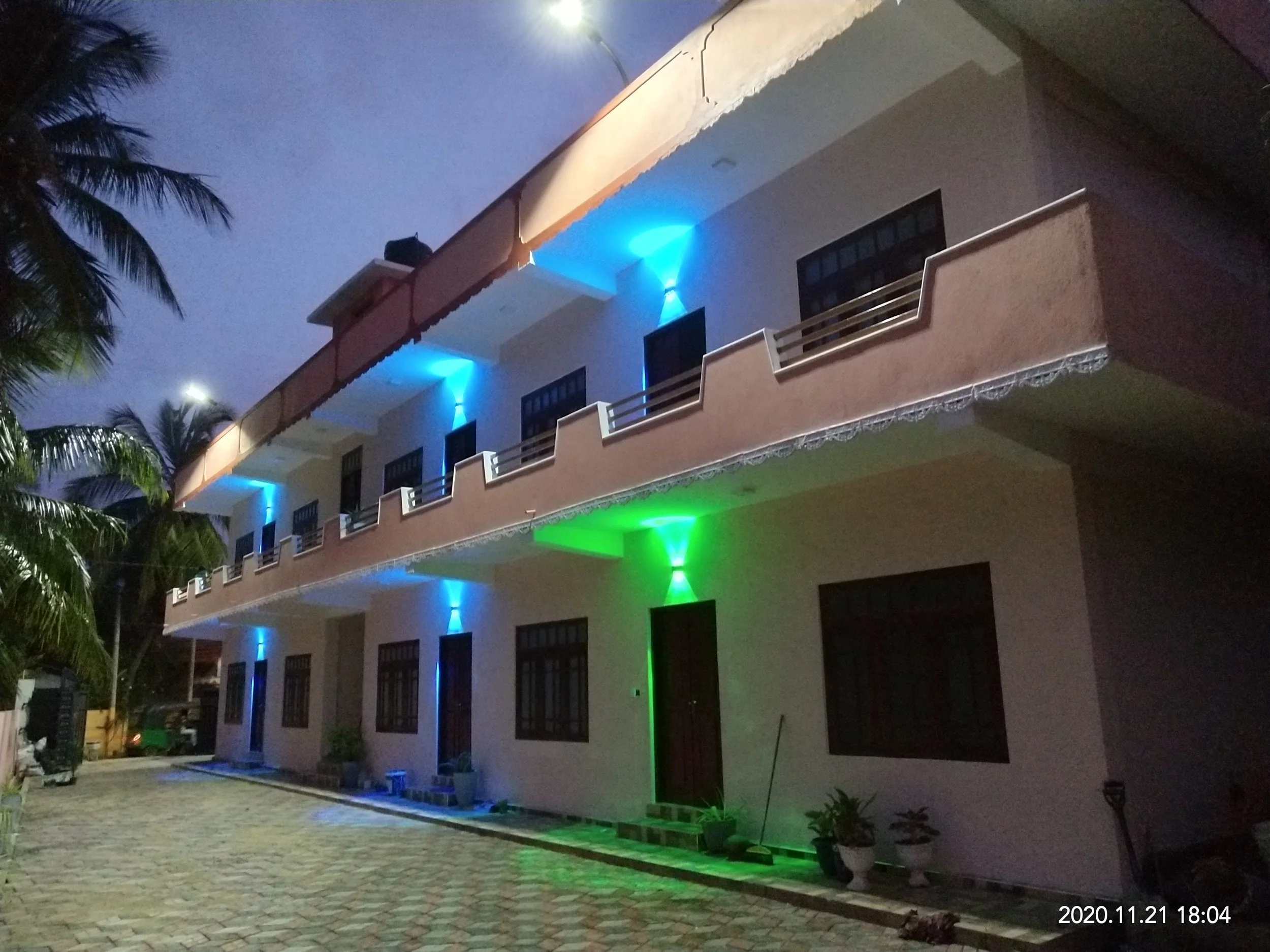 A multi-story building at night with exterior lighting in green and blue. The building has balconies and several windows. There are palm trees nearby, and a paved area in the foreground. The date and time stamp in the bottom right corner shows '2020.