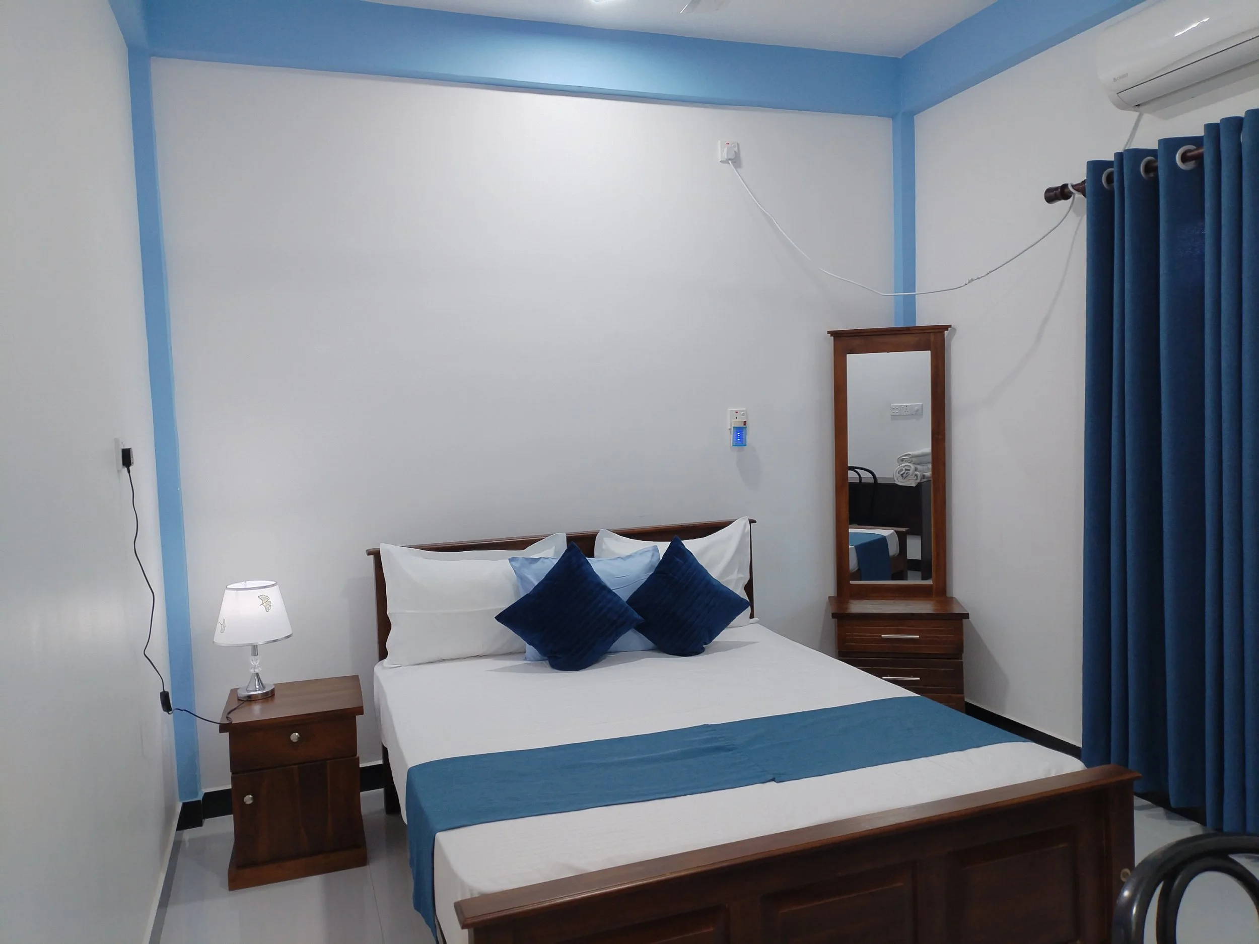 A bedroom with a bed, bedside table, and a mirror. The room has white walls with blue accents on the ceilings, window frame, and curtains. An air conditioner is mounted on the wall.