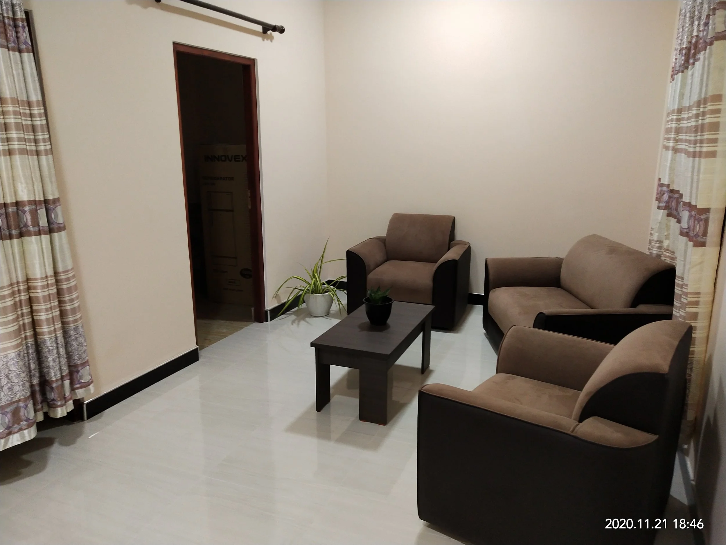 A living room with beige walls, a brown armchair, a sofa, a small black coffee table with potted plants, and curtains with patterned stripes. There is a doorway leading to another room.