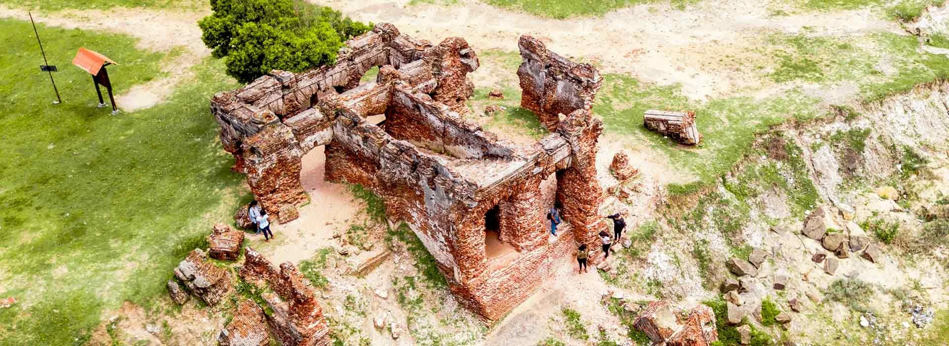 Aerial view of ruins of an ancient brick building with several visitors examining the site, surrounded by grassy and rocky terrain.
