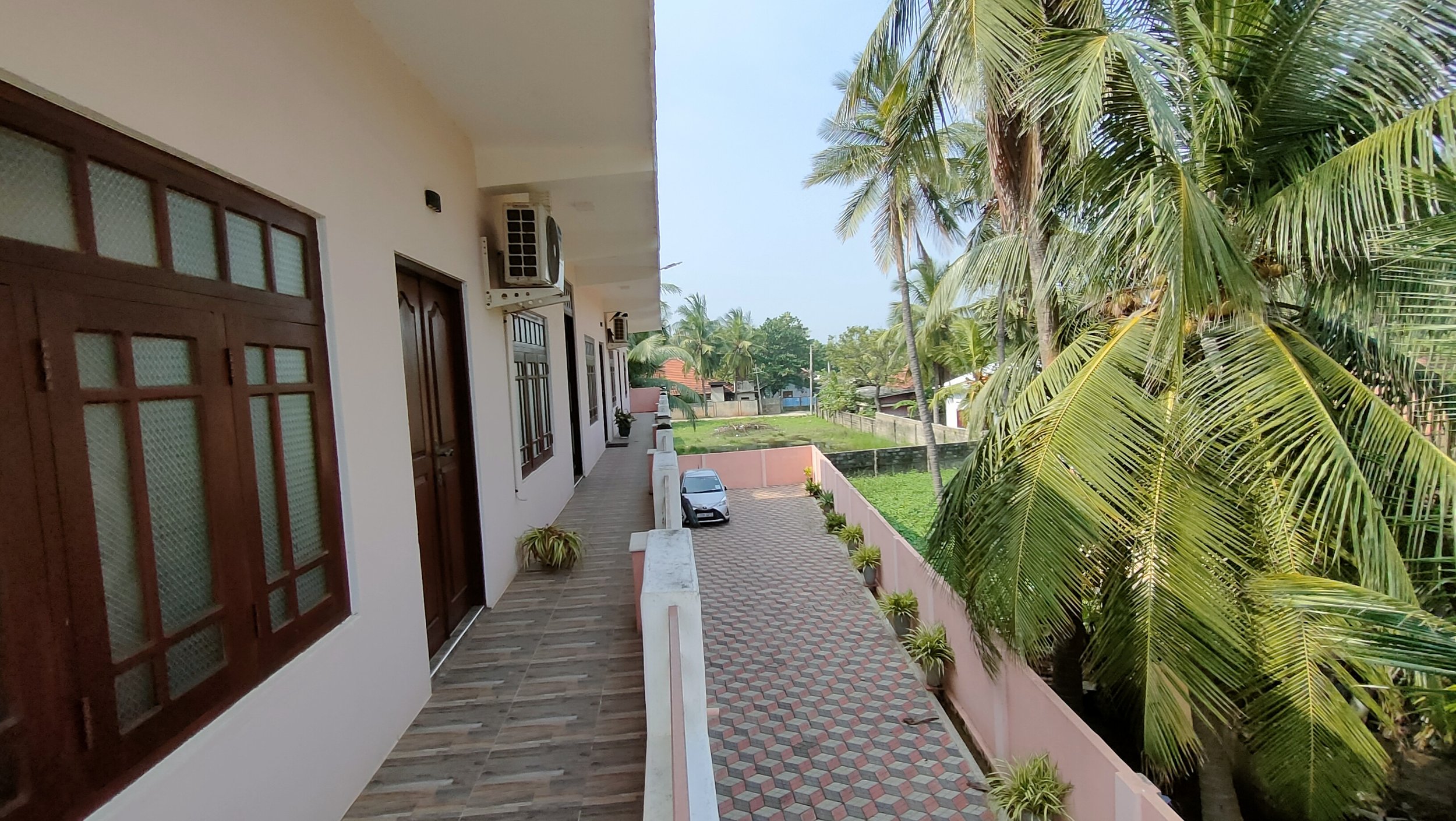 Balcony view with brown door and window on white building, tropical palm trees, parked silver car, green grass, pink boundary wall, and colorful rooftops in the background.