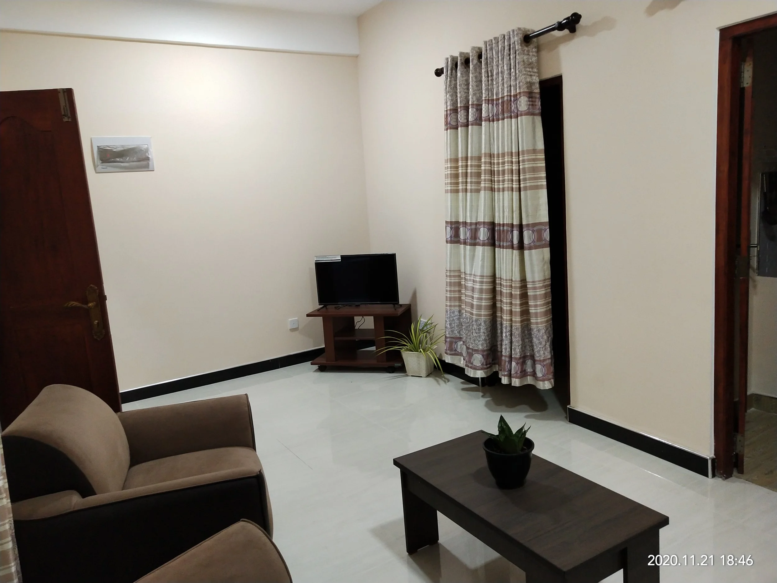 Living room with a couch, coffee table, small TV on a stand, window with curtains, potted plant, and a door frame.