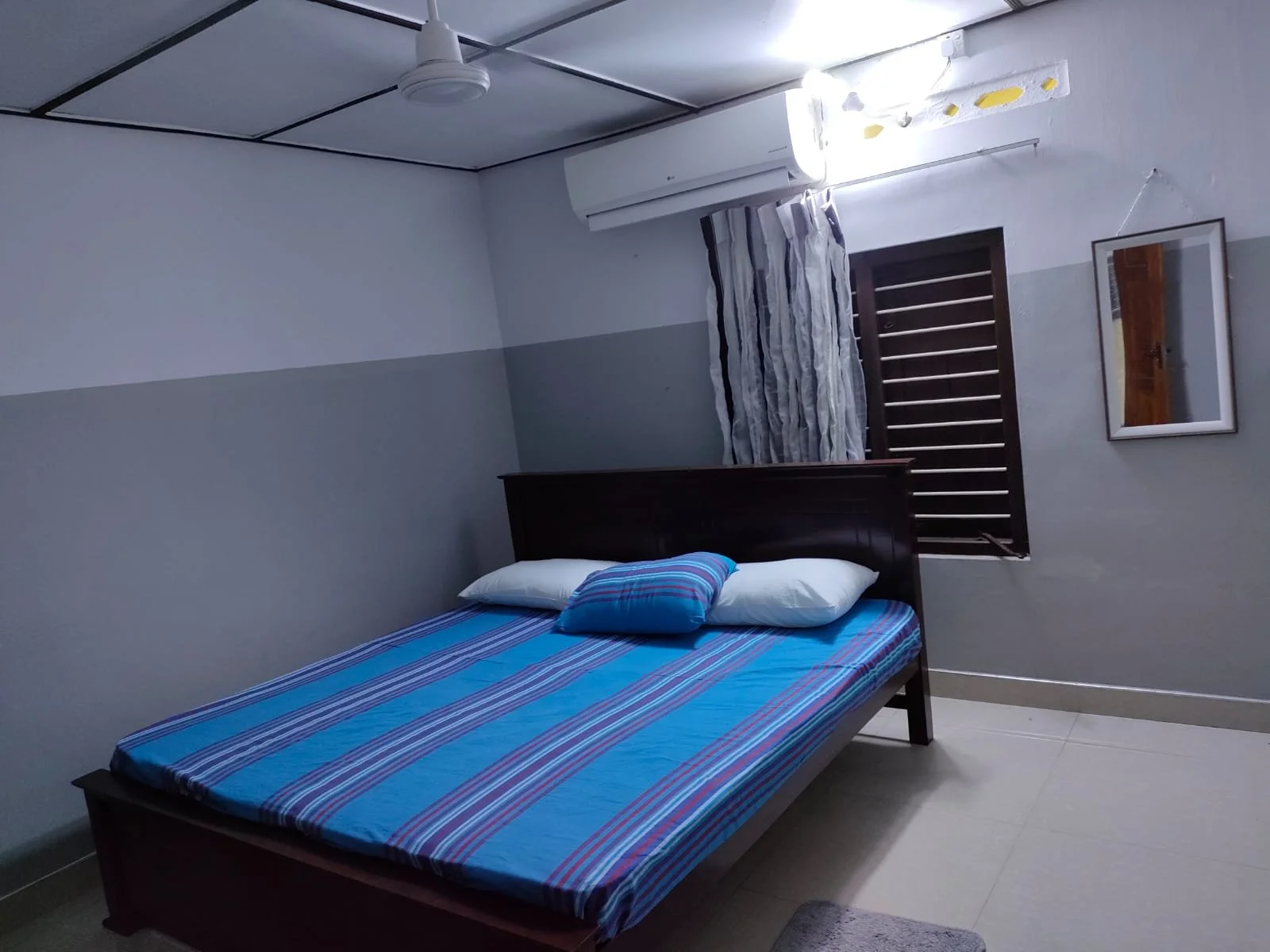 A bedroom with a wooden bed frame, a blue striped mattress, three pillows, a window with striped curtains, a wall mirror, a ceiling fan, and an air conditioning unit.