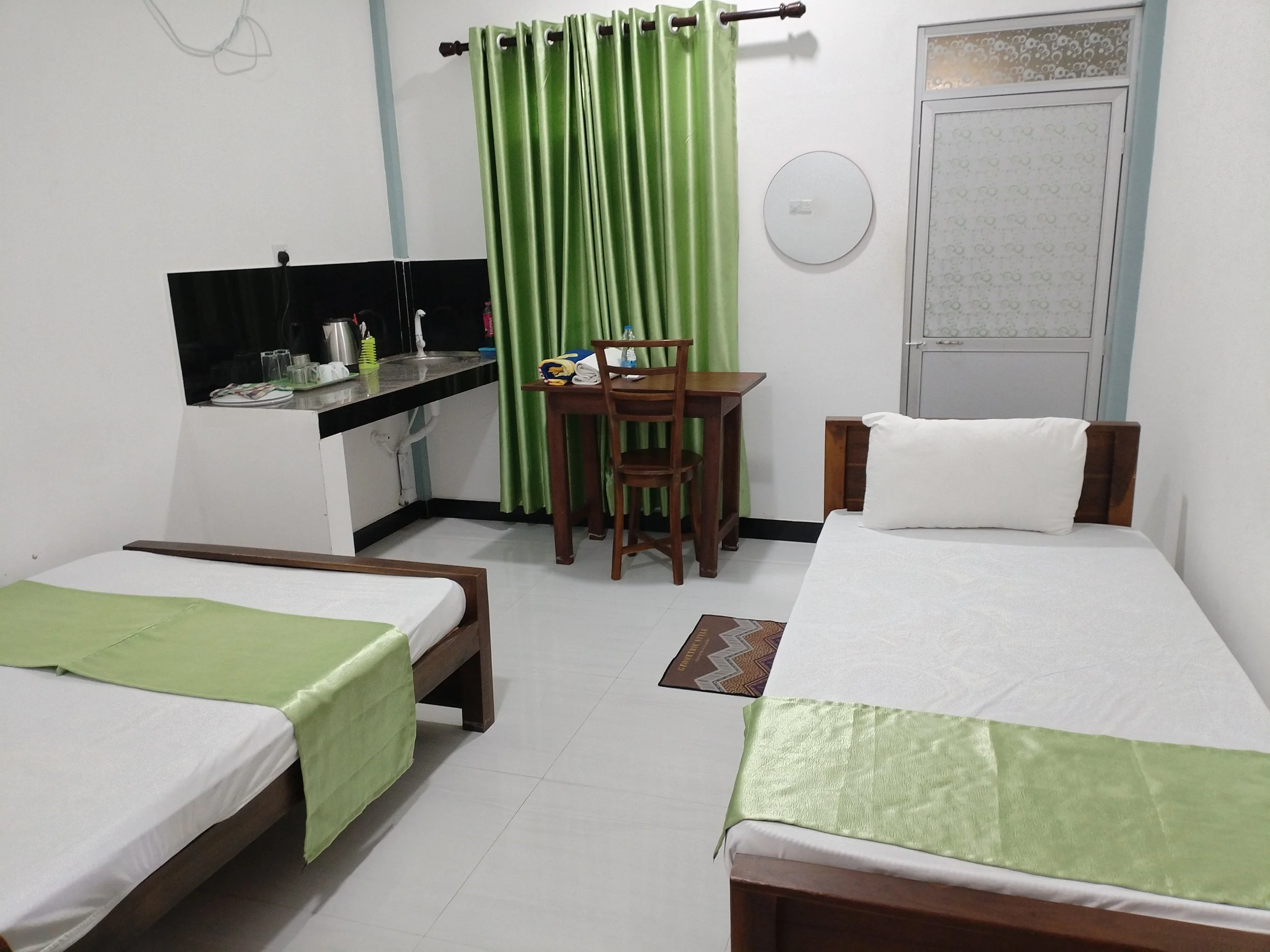A room with three beds, each covered with light green bed sheets, and two connected twin beds visible in the foreground, with a small wooden table and chairs, a kitchen countertop with utensils, a sink, a water bottle, and a window with green curtain