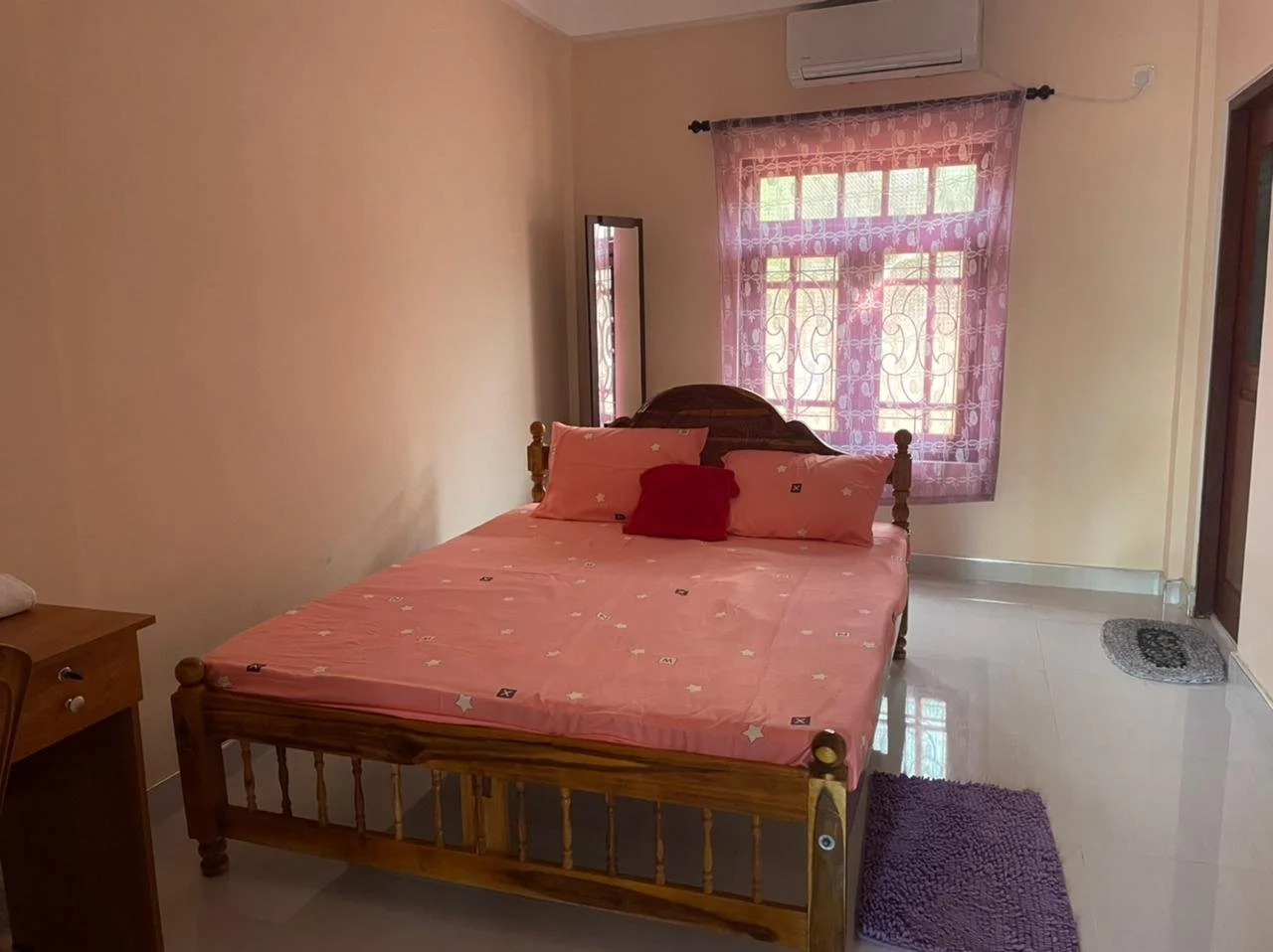 A bedroom with a wooden bed, pink bedding, and pillows, pink curtains, and a mirror, with a small wooden desk and a chair nearby.
