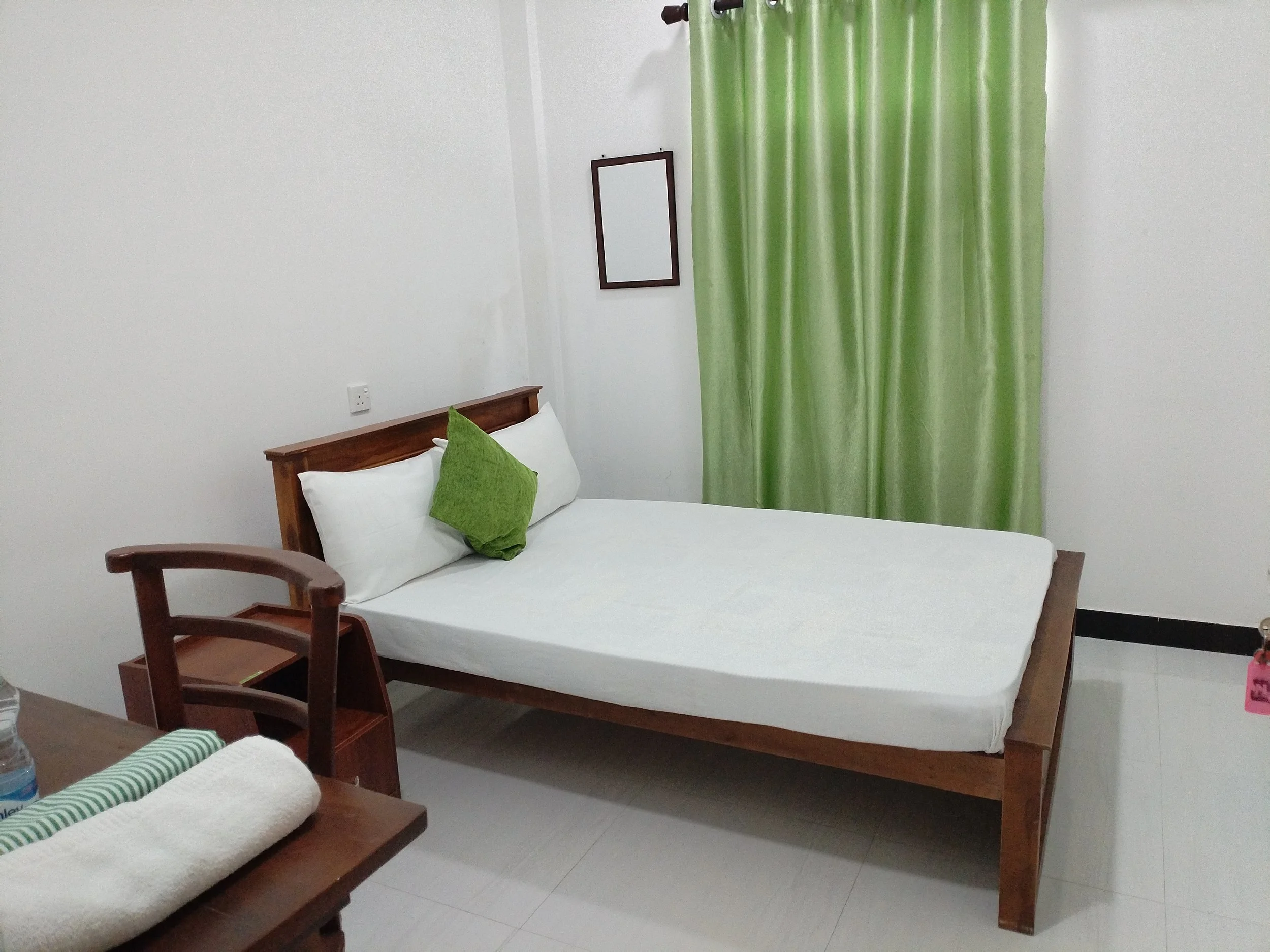 A bedroom with white walls, a wooden bed frame, and green curtains. The bed has white and green pillows. A small framed mirror hangs on the wall, and there's an electrical outlet near the head of the bed. Part of a wooden chair and a table with a rol