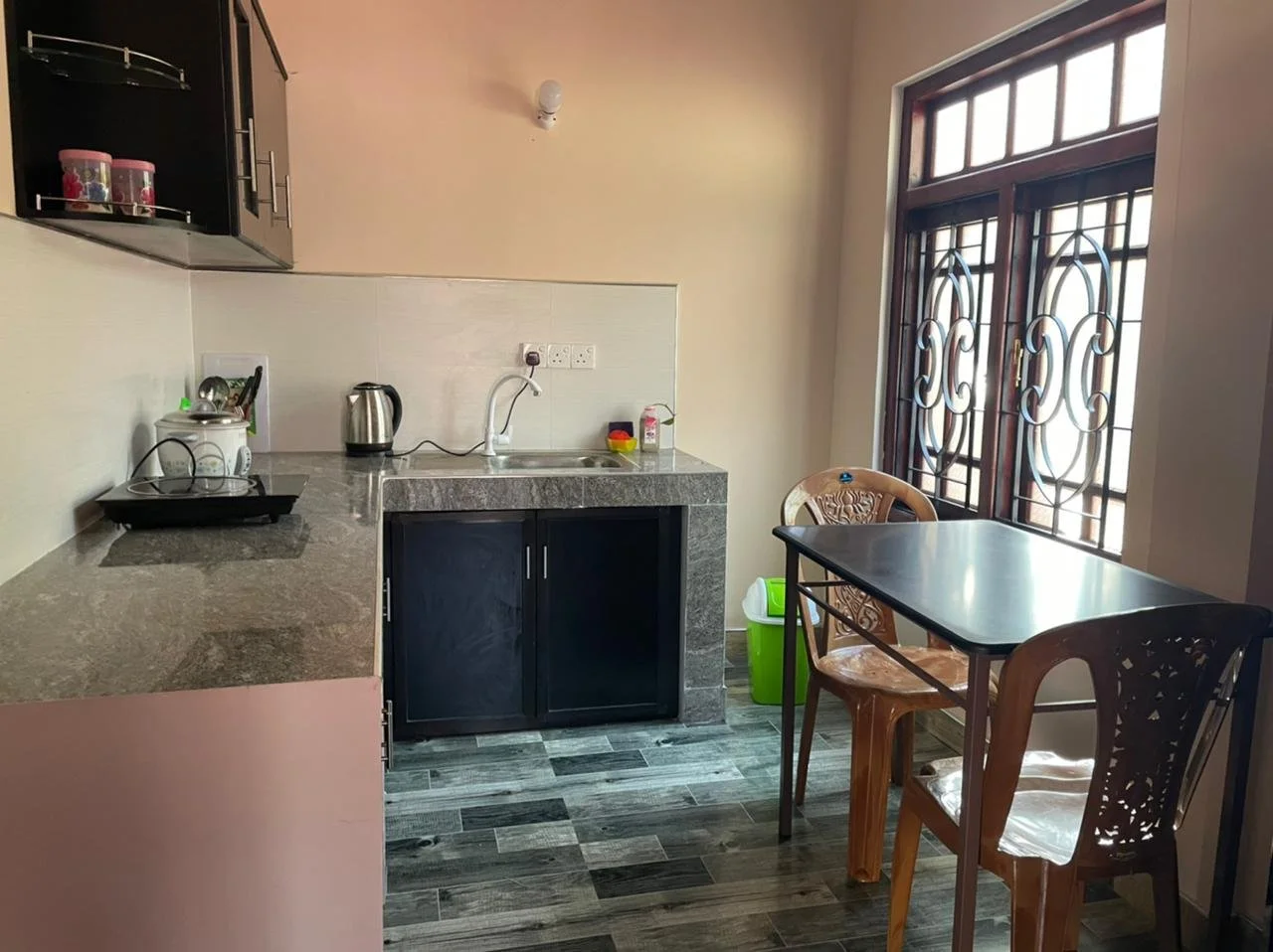 A small kitchen with a gray granite countertop, black cabinets, a stainless steel kettle, a white rice cooker, and a black electric stove. There is a double electrical outlet above the countertop. A large window with decorative iron bars lets in natu