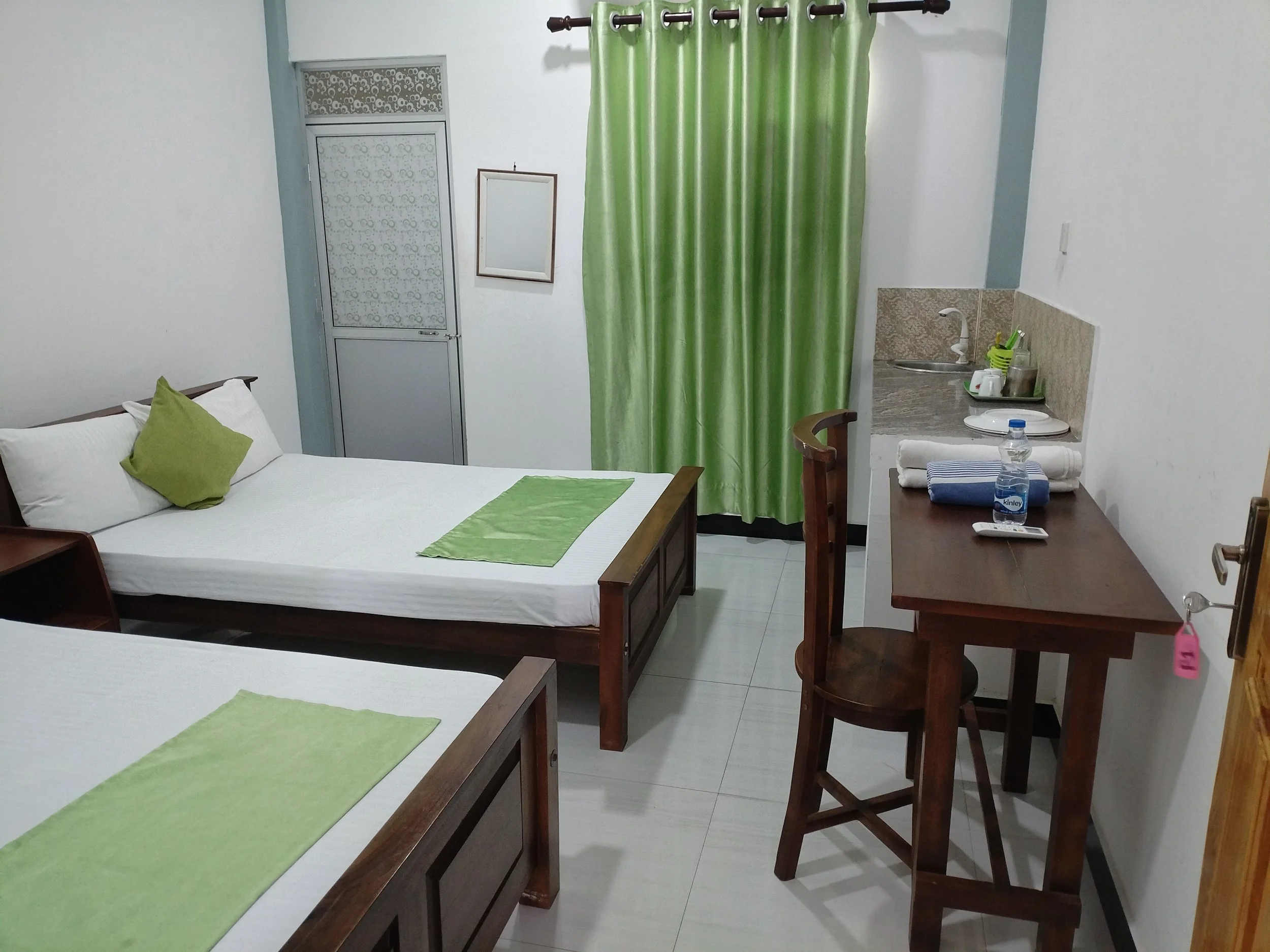 A cozy bedroom with two neatly made beds, each with white bedding and green accent pillows, a small wooden table with towels and a water bottle, a kitchenette with a sink and utensils, a mirror, and a window covered with a floral-patterned curtain.