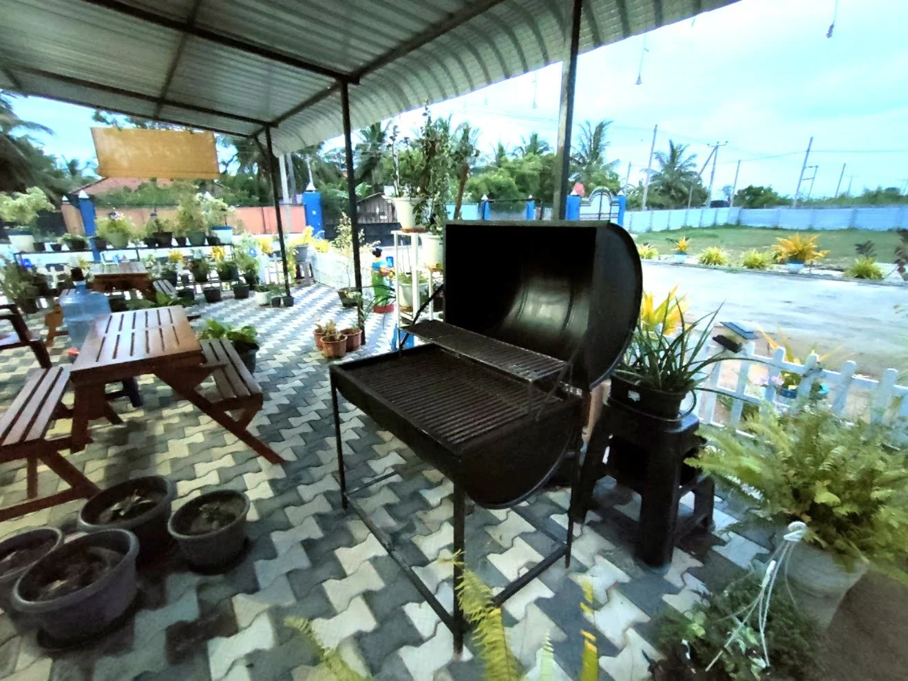 Outdoor patio area with a covered roof, potted plants, wooden benches, a black barbecue grill, and a decorative tiled floor, overlooking a green landscape with trees and a white fence.