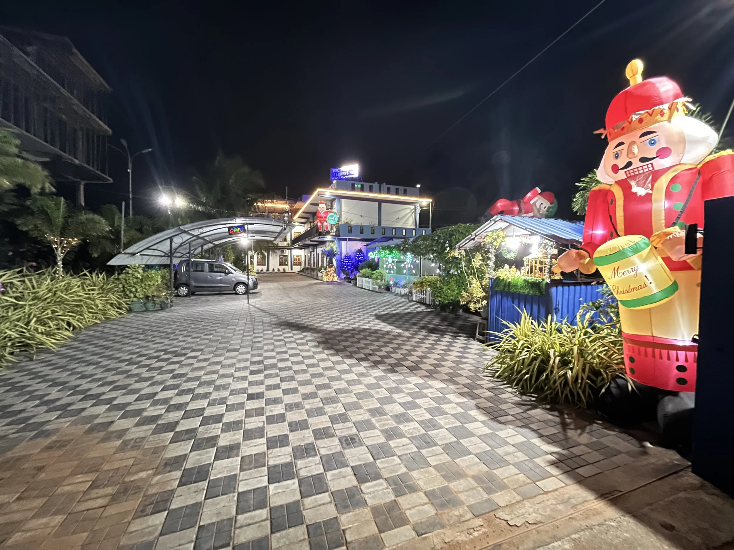 A night scene of a decorated outdoor area with inflatable Christmas figures, including a large Santa Claus and a nutcracker, illuminated with colorful lights, next to a building and parked cars.