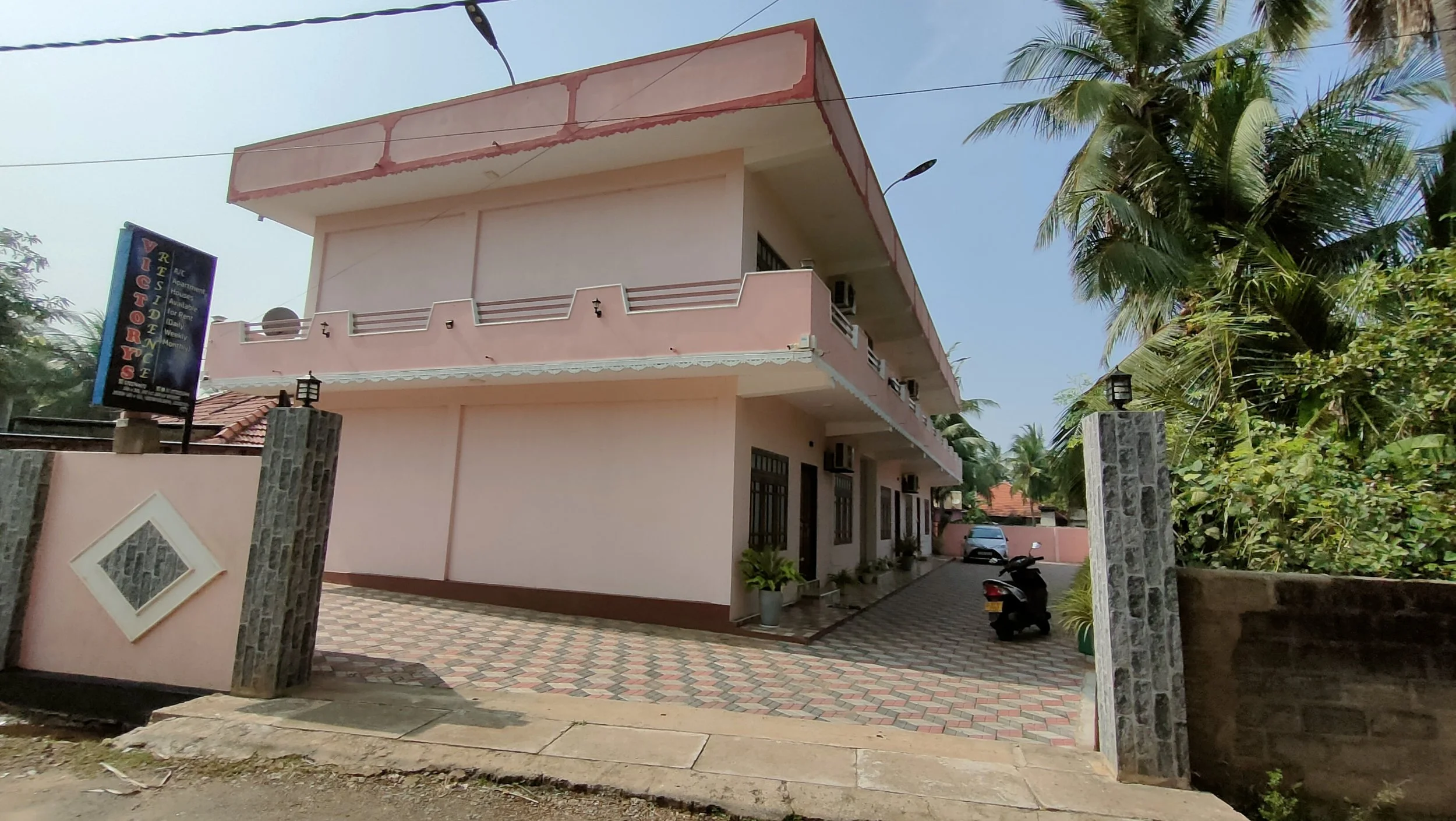 A pink multi-story building with a small courtyard, surrounded by palm trees and tropical plants, with a parked scooter and a car in the background.