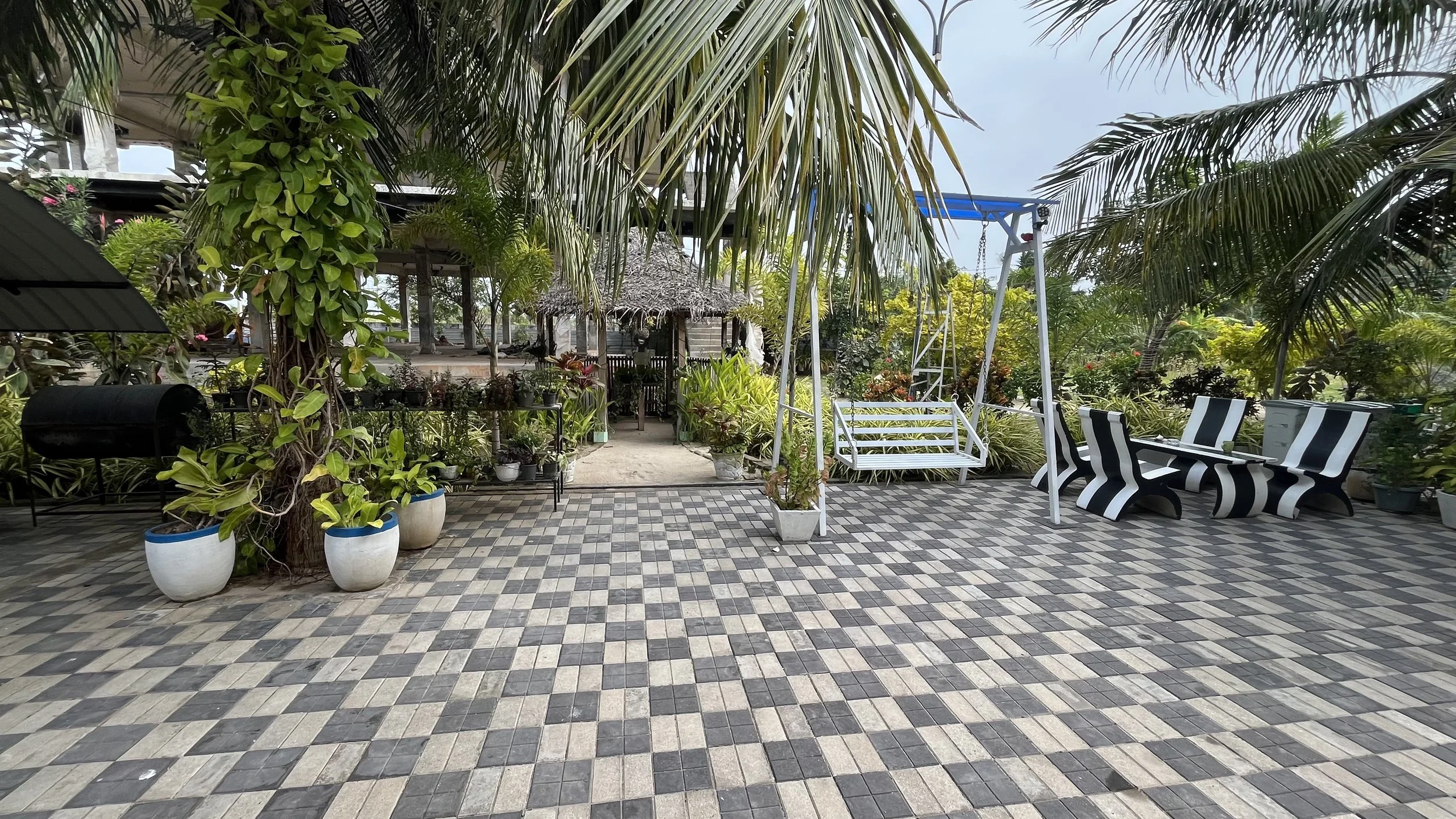 Outdoor patio with black and white striped benches, potted plants, palm trees, a swing, and thatched-roof hut in the background.
