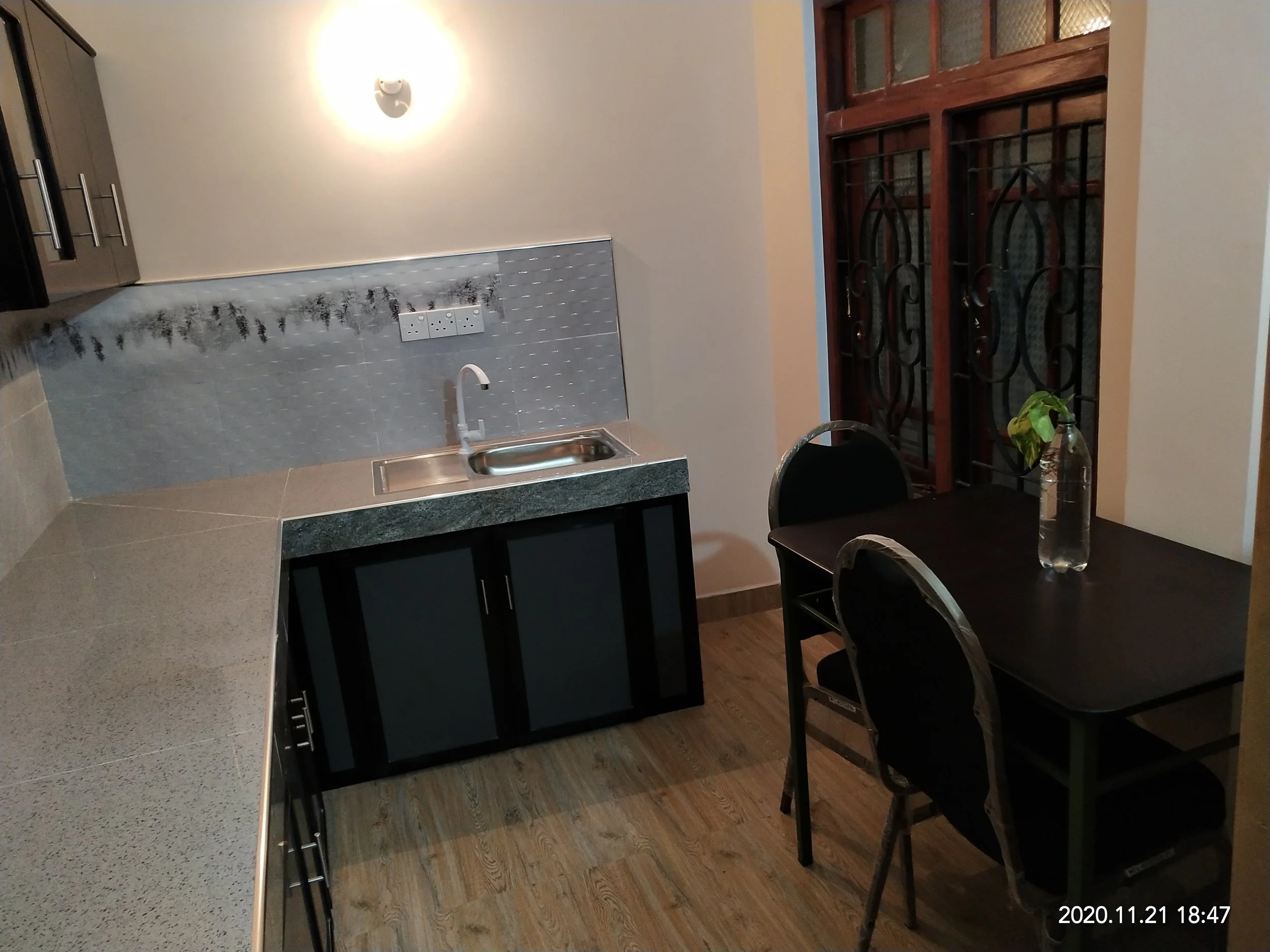A kitchen and dining area with a gray countertop, a stainless steel sink, a black chair, a wooden window with decorative black metal bars, a table with a water bottle and green plant, and a wall-mounted light fixture.