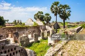 Ruins of old stone buildings with palm trees and clear blue sky in the background.