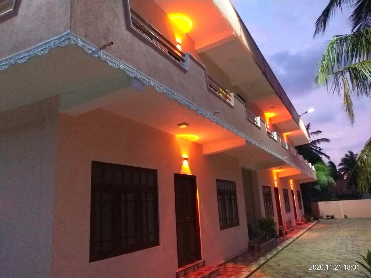 Two-story building outdoors at dusk with orange wall-mounted lights on the balcony and exterior walls, surrounded by trees and a paved courtyard.