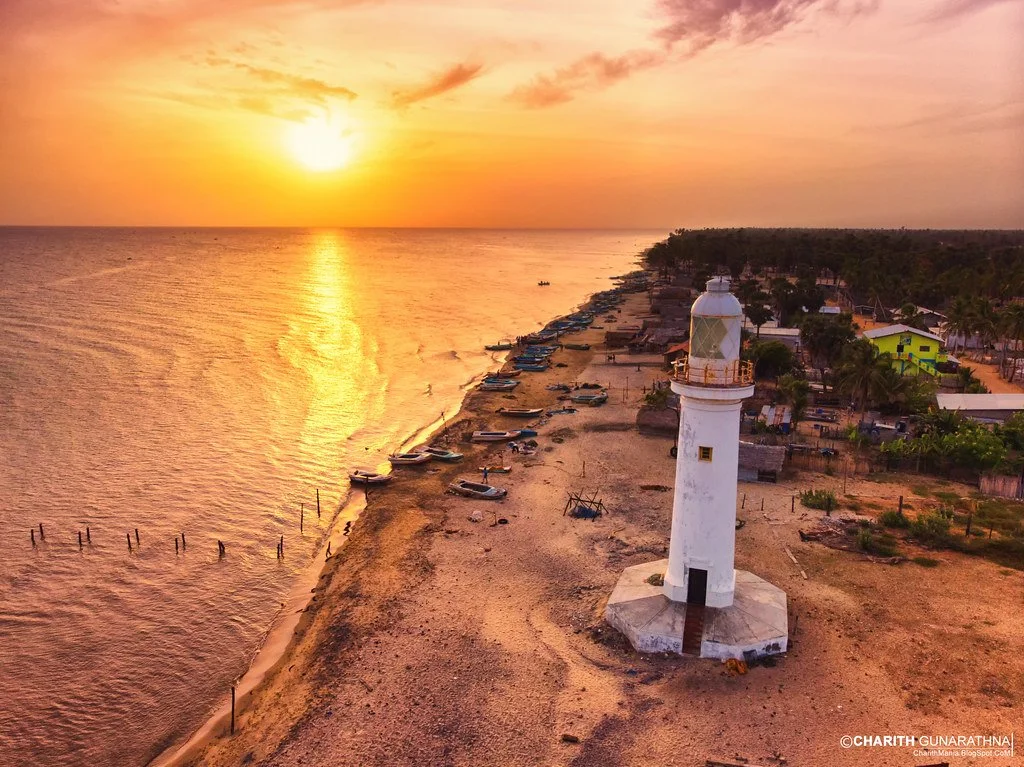 Sunset over a coastal area with boats, a lighthouse, and trees.