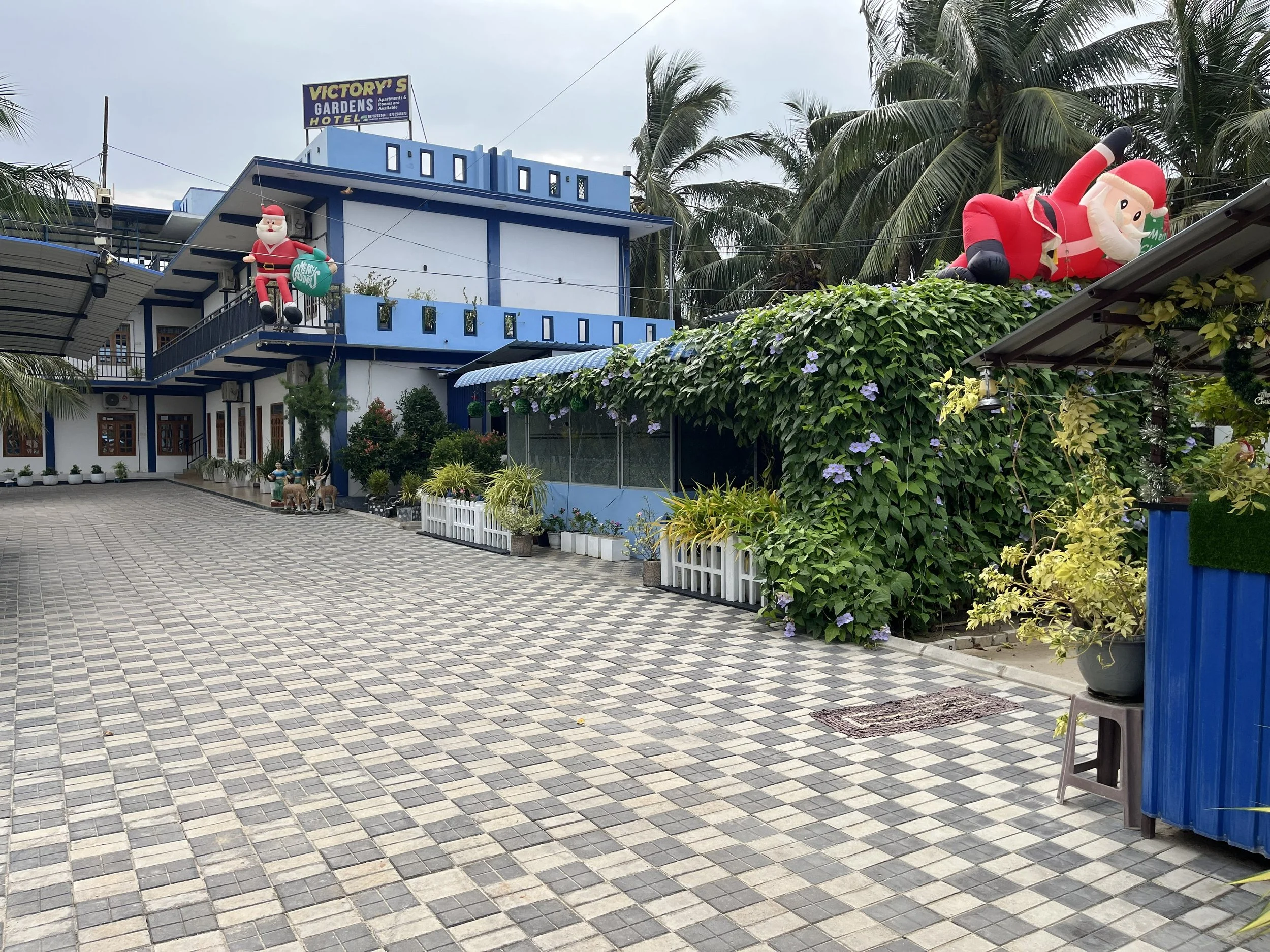 Inflatable Santa Claus and inflatable Santa figure hanging on a building surrounded by green foliage and palm trees, with a tiled courtyard in the foreground.