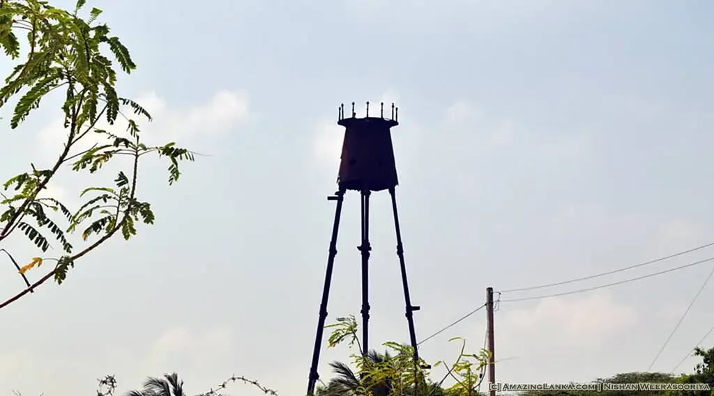Tall, black water tower with an approaching sunny sky, surrounded by green trees and plants.