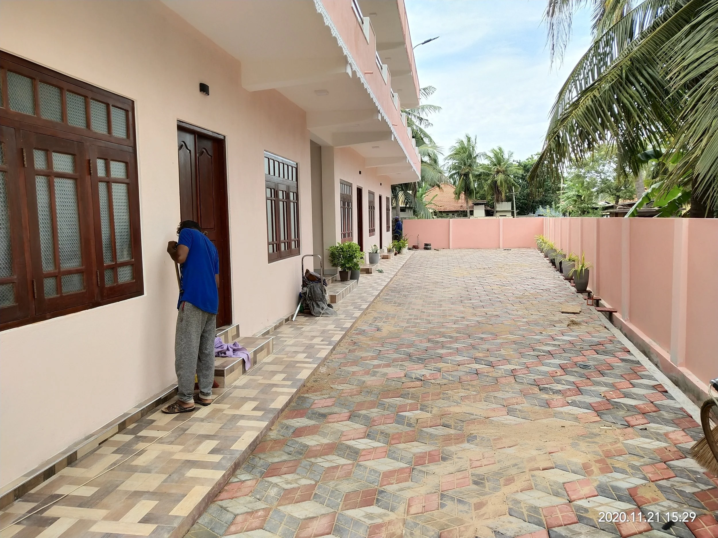 A man in a blue shirt and gray sweatpants sweeping the outdoor tiled patio area with a broom.