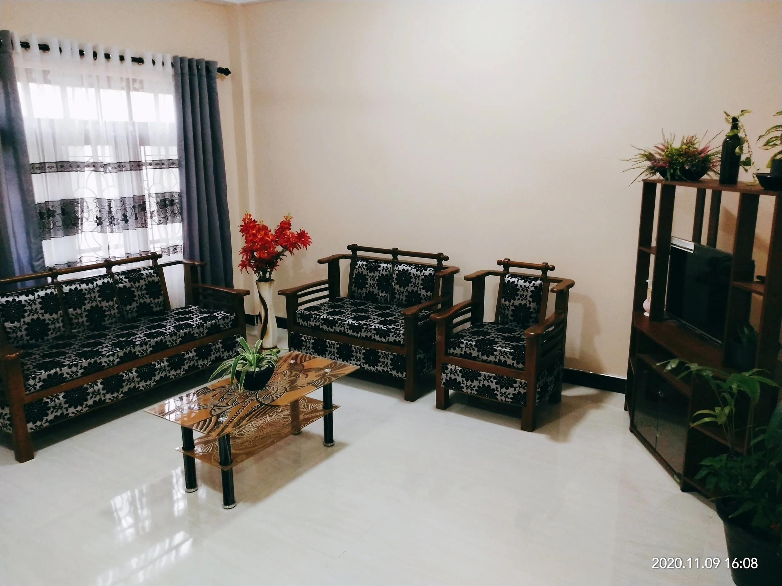 A living room with beige walls, a window with white curtains and blue drapes, and furniture that includes a wooden sofa and chairs with black and white floral upholstery. There is a small wooden coffee table with a leaf-patterned tile top, a vase wit