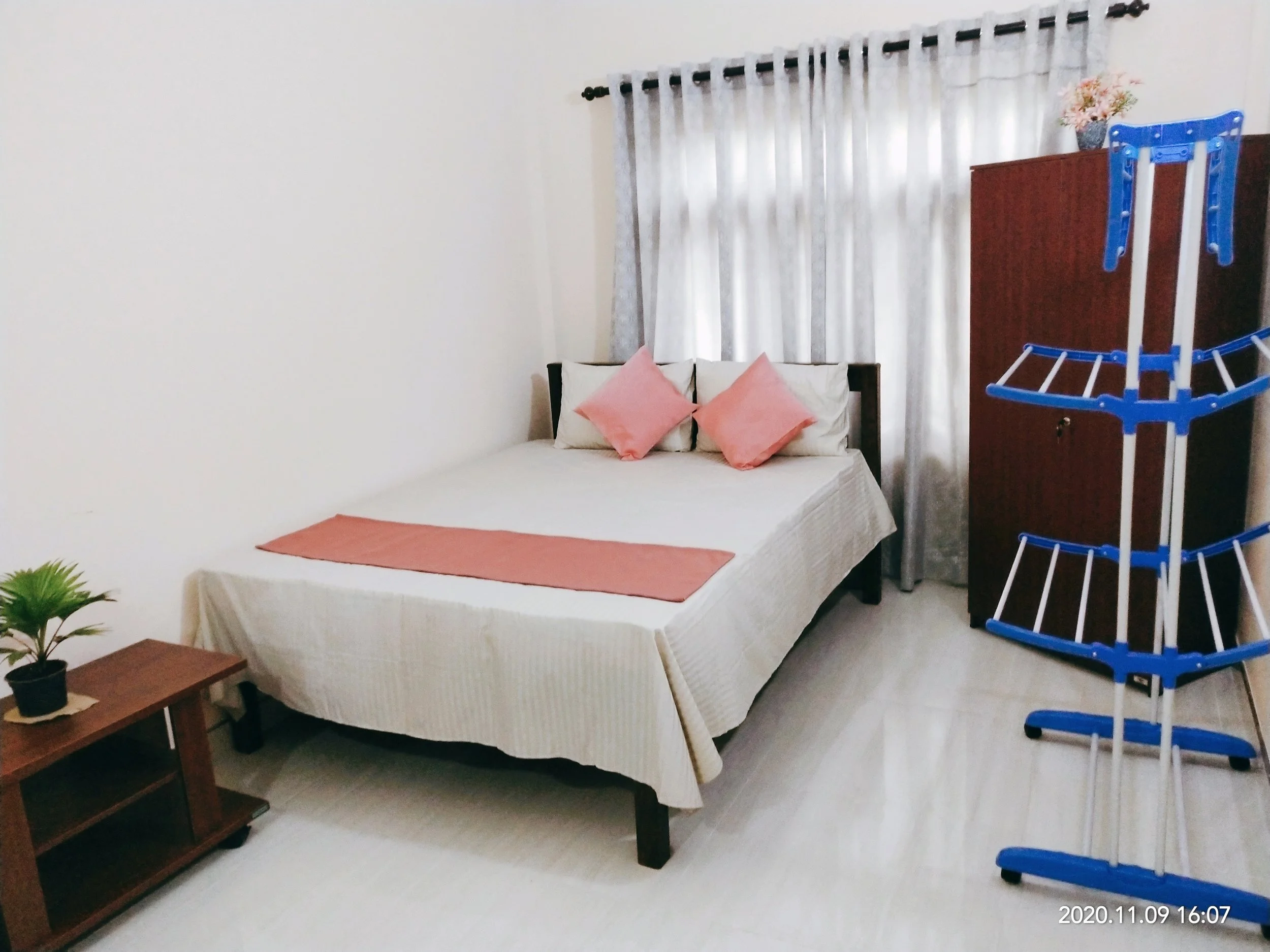 A bedroom with a bed covered in white linens and pink pillows, a wooden nightstand with a small potted plant, futon or daybed, and a wooden wardrobe with a flower vase on top. There is a blue drying rack in the room and white curtains allowing light through the window.